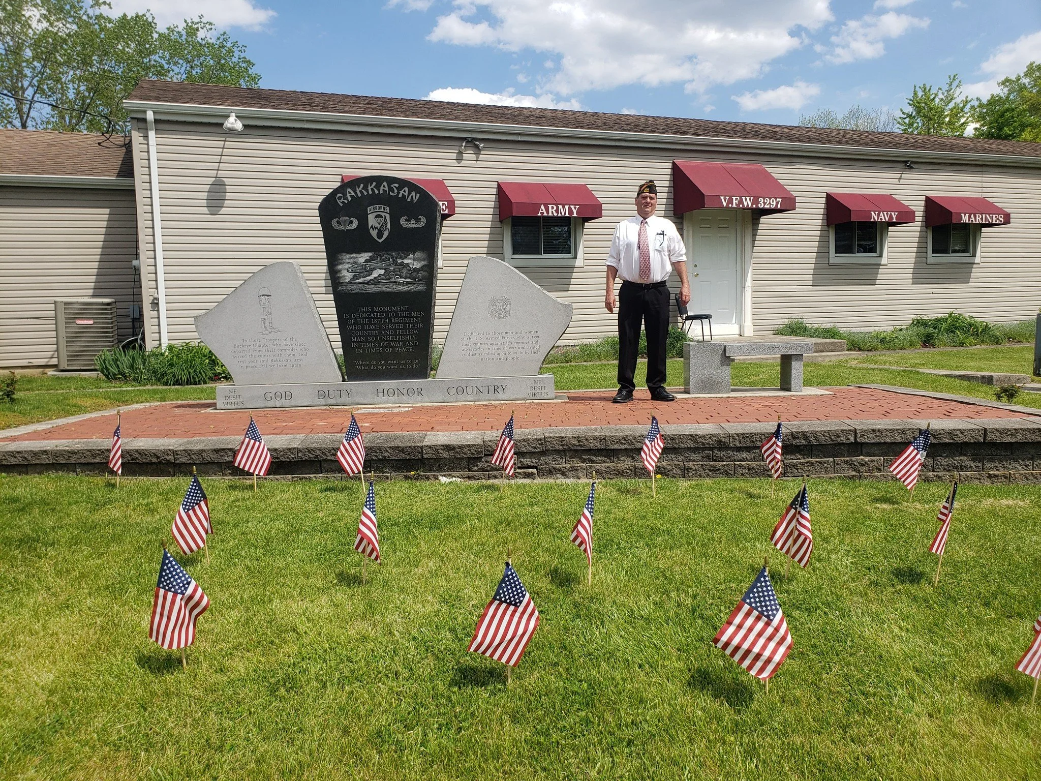 A memorial display dedicated to the armed forces with a large black and gray stone monument and three smaller gray stone plaques, surrounded by small American flags on a grassy lawn. A man dressed in a white shirt, black pants, and a tie stands beside the memorial. In the background, there is a beige building with red awnings labeled 'ARMY,' 'V.F.W. 3297,' 'NAVY,' and 'MARINES.'