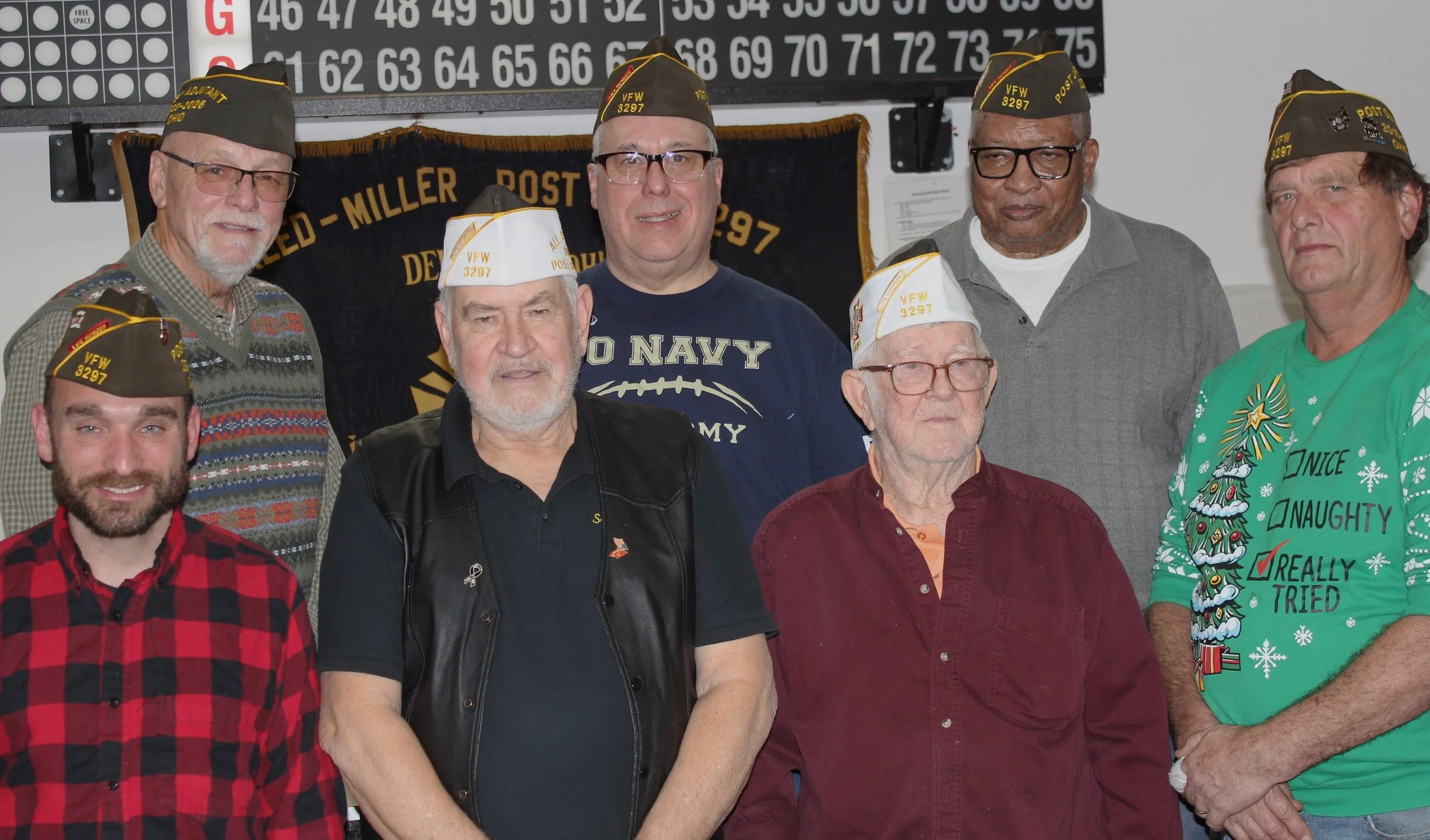 Group of eight men, some wearing military veterans hats, gathered indoors in front of a Bingo board and a black banner with gold text. Some men are dressed casually, with one wearing a Christmas-themed shirt.