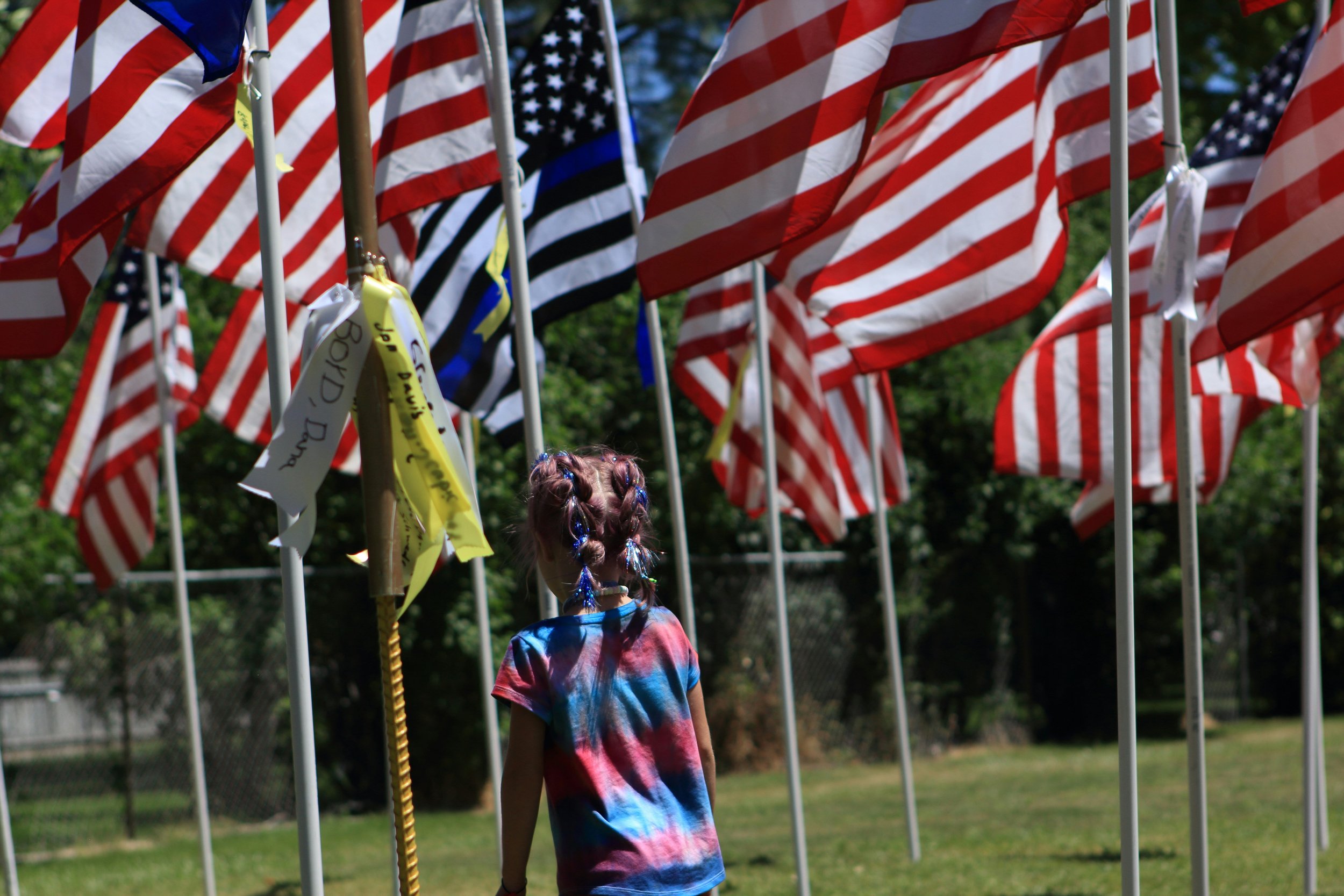 A young girl with braided hair, wearing a tie-dye shirt, looking at a display of American flags on flagpoles outdoors.