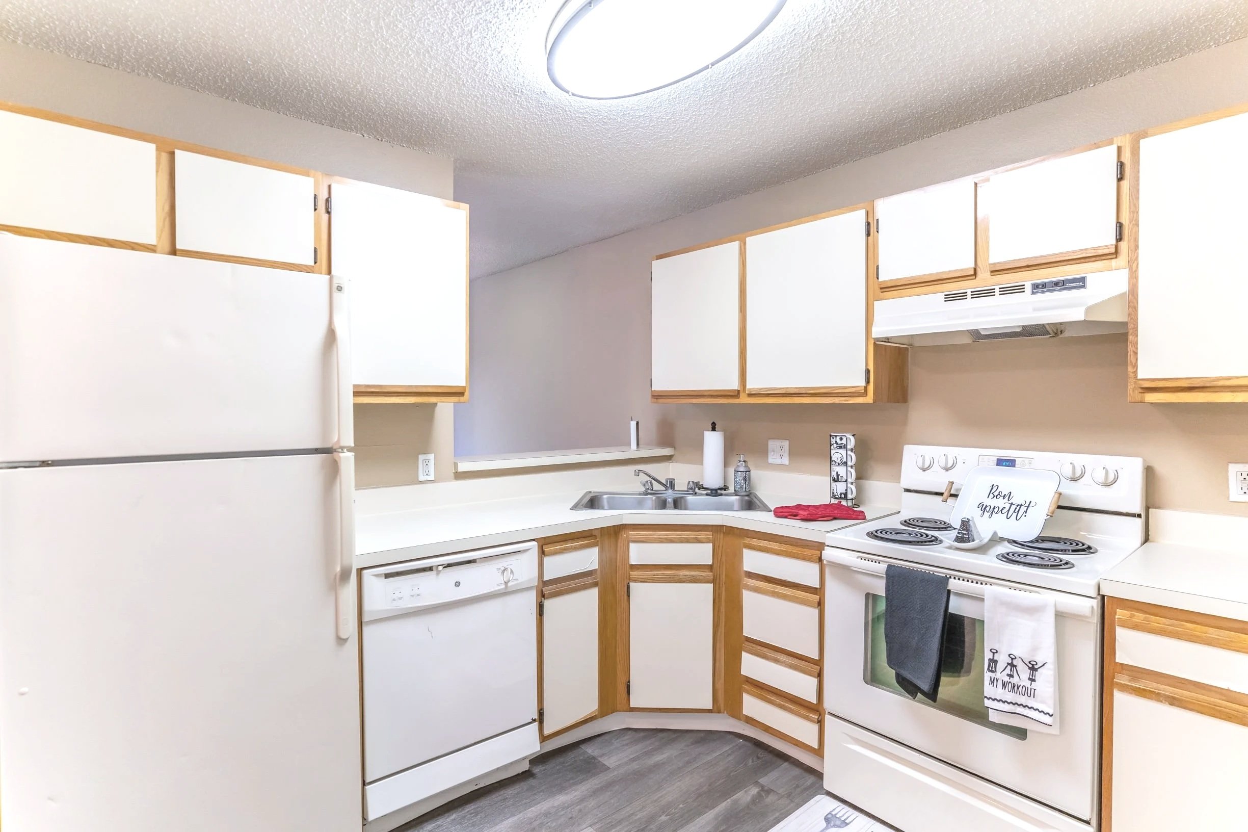 Kitchen with white appliances and wooden cabinets, including refrigerator, dishwasher, and stove, with a corner double sink and gray flooring.