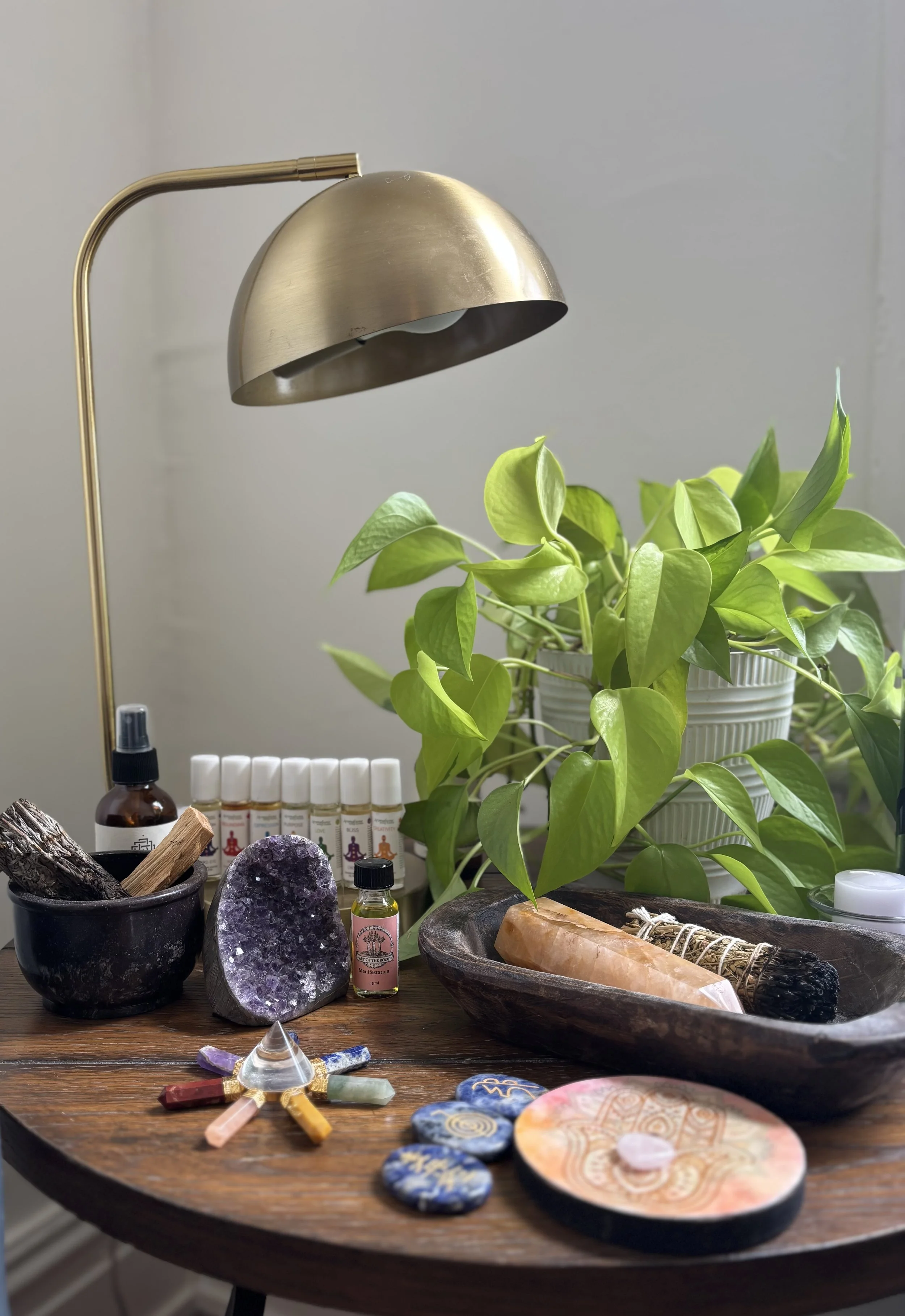 A wooden table holds a potted green plant, a large purple geode, small bottles, crystals, a tarot card layout, and a wooden bowl with a salt lamp and a smudge stick, illuminated by a brass desk lamp.
