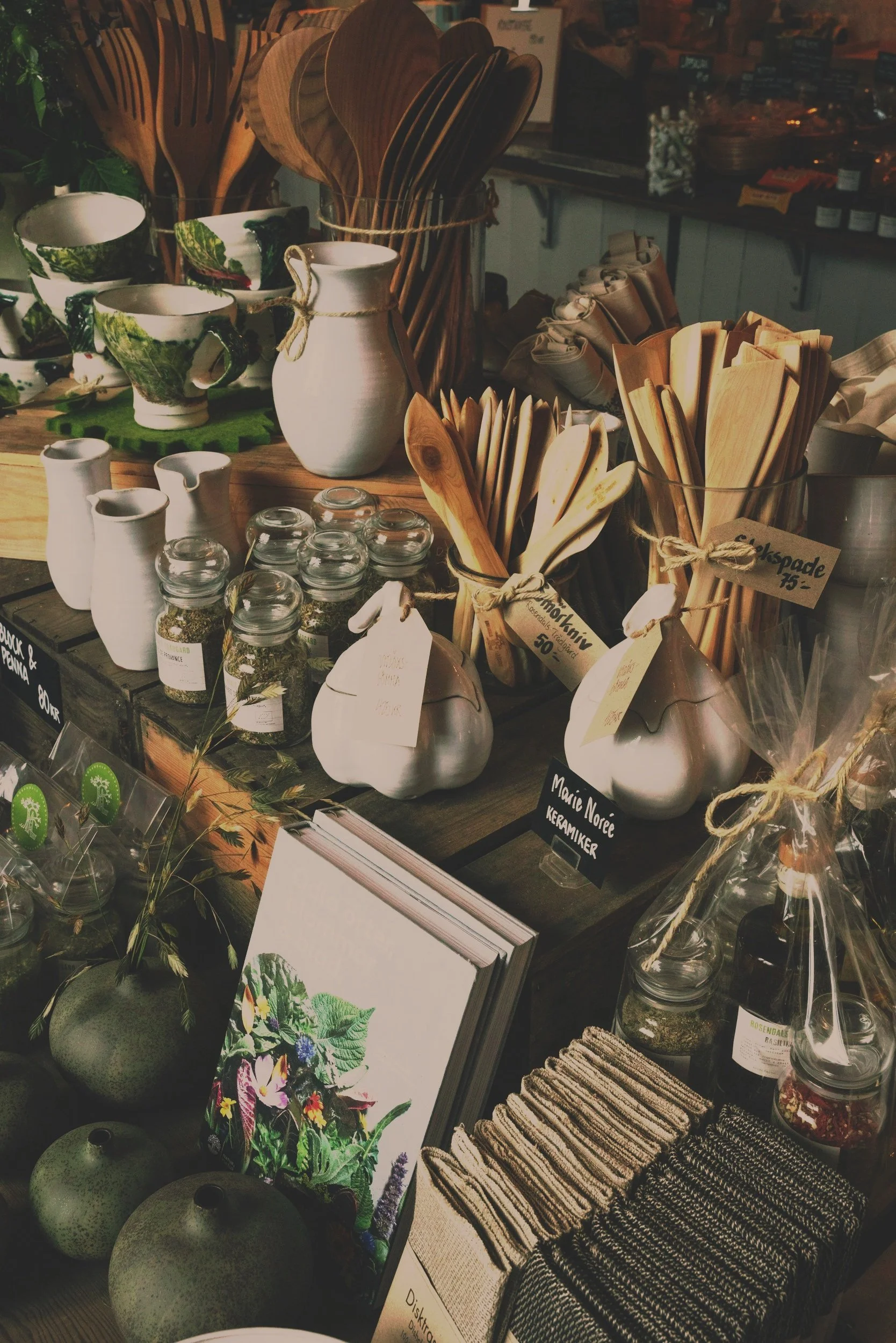 A display of ceramic and wooden kitchenware, including bowls, cups, and utensils, with small jars of herbs, decorative gourds, a framed botanical illustration, and cloth napkins on a wooden table in a shop. market wares goods