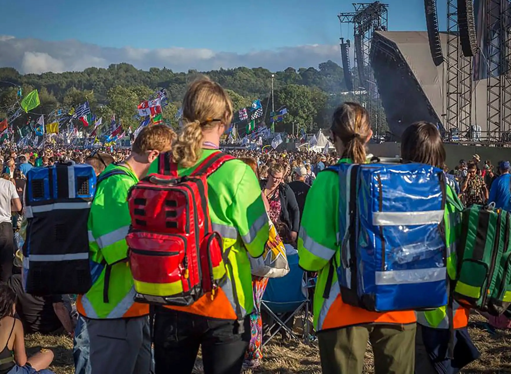 Four emergency responders wearing bright green jackets with reflective stripes and colorful backpacks, standing in front of a large crowd at an outdoor festival with a stage, flags, and trees in the background.
