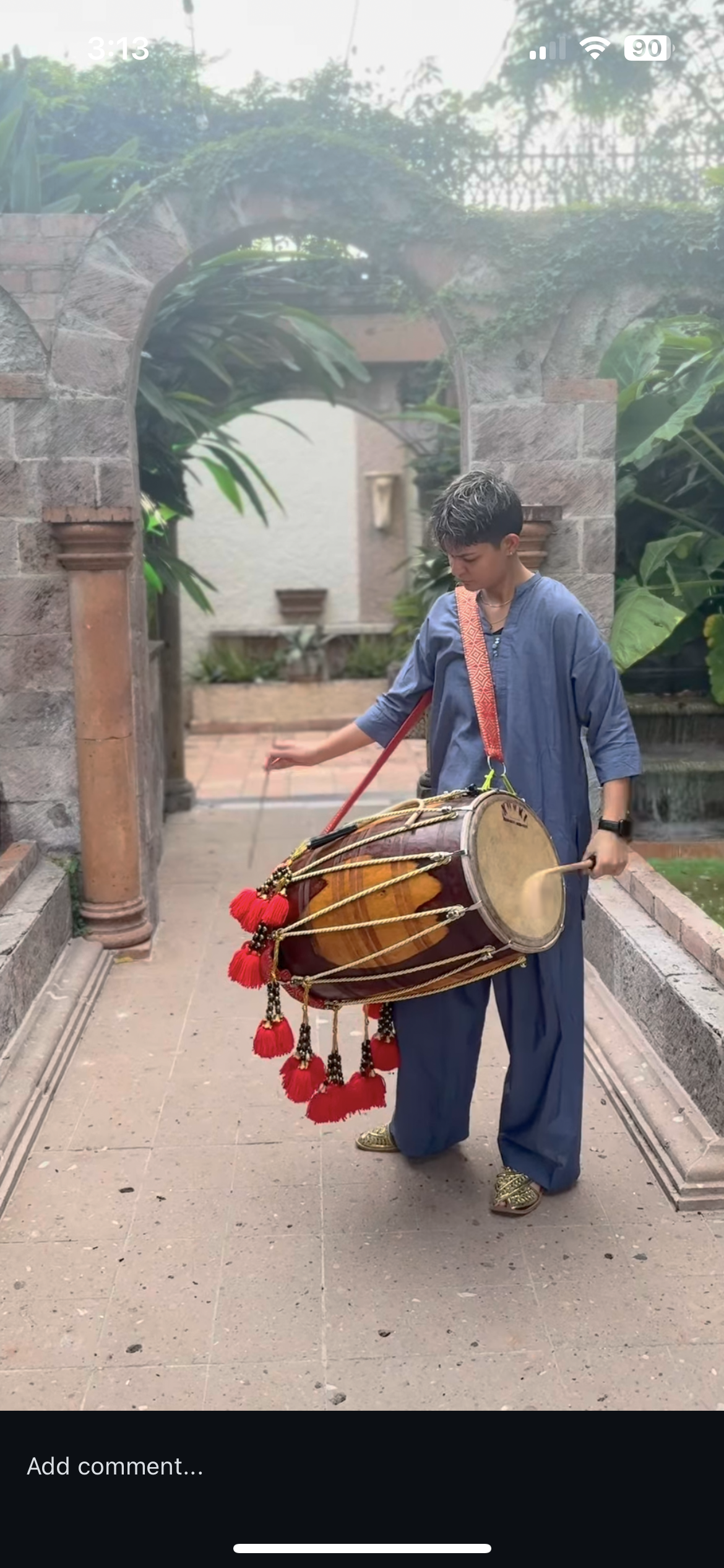 A young woman dressed in blue traditional clothing playing a large indian // pakistani dhol drum decorated with red tassels outdoors.