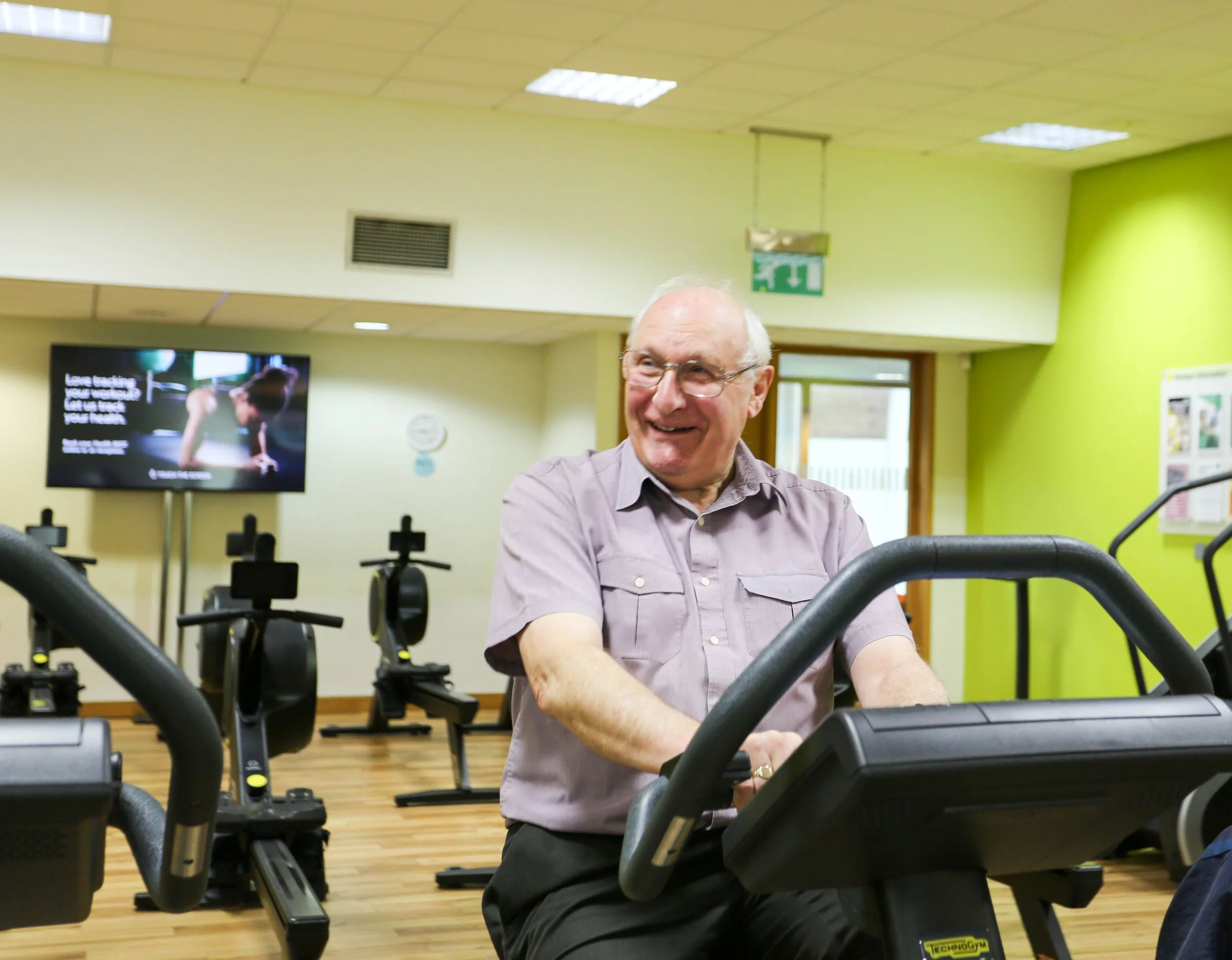 An older man smiling while seated on exercise equipment in a gym with green walls and a television in the background.
