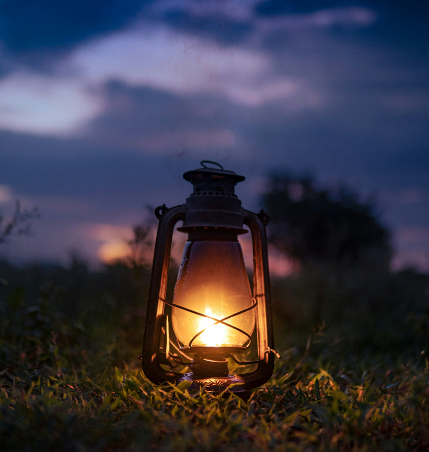 An old-fashioned oil lantern with a lit flame, placed on grass outdoors at dusk with a dark sky and cloudy background.