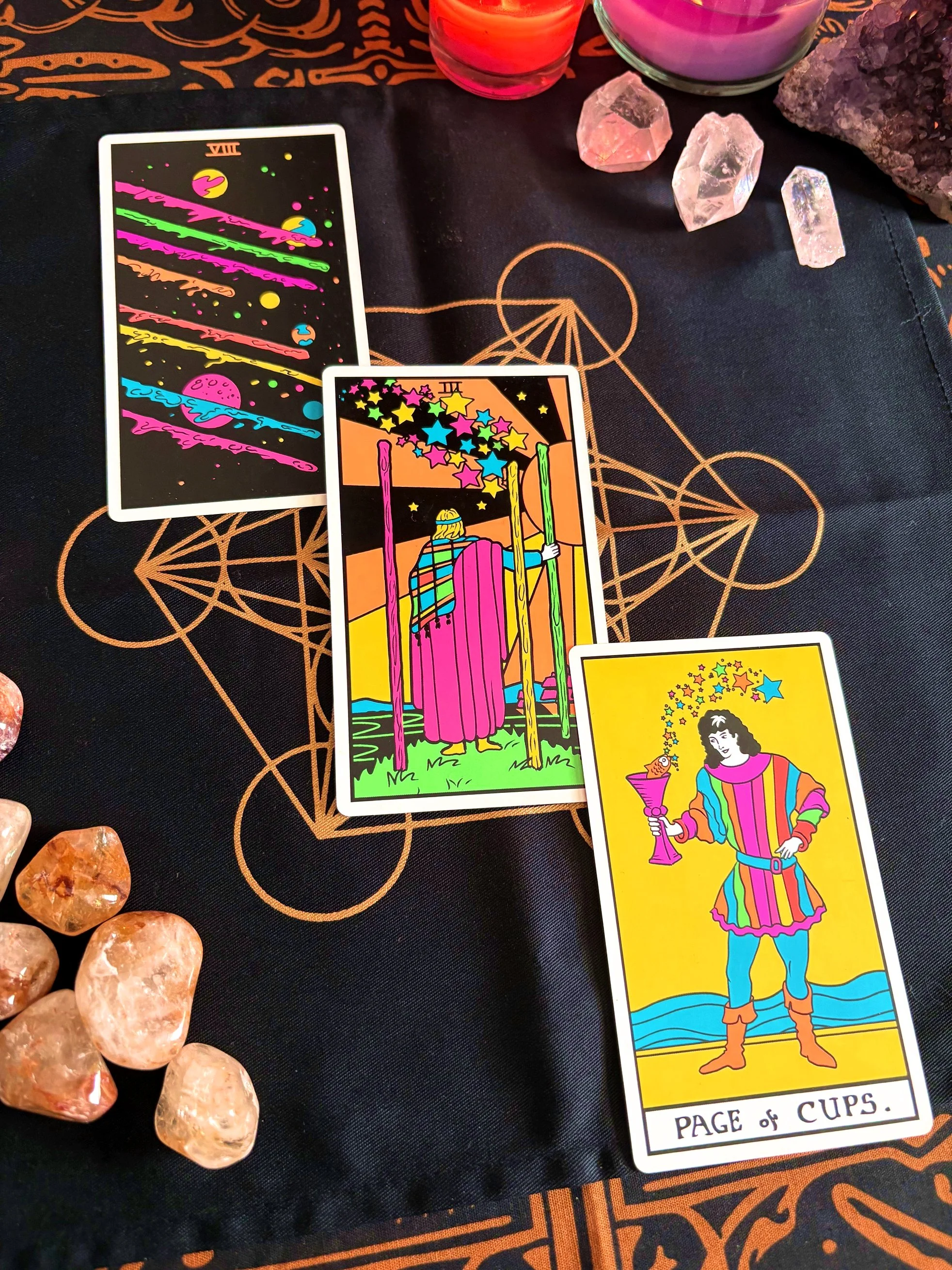 Colorful tarot cards laid out on a black cloth with geometric and mystical patterns, surrounded by rose quartz and other crystals, with candles in the background.