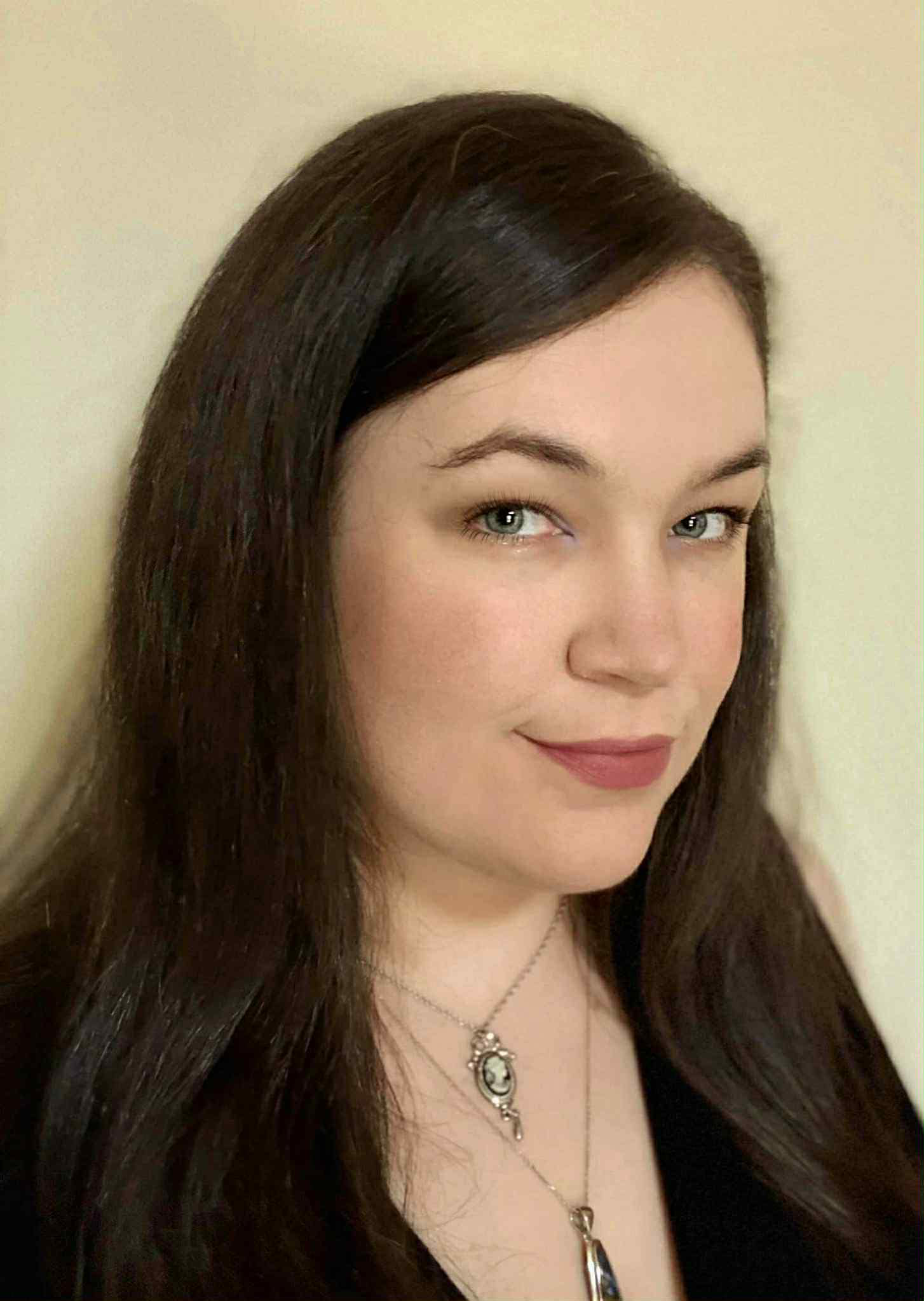 A young woman with long dark hair, light-colored eyes, and fair skin looking at the camera. She has a slight smile and is wearing a necklace with a pendant. The background is plain and light-colored.