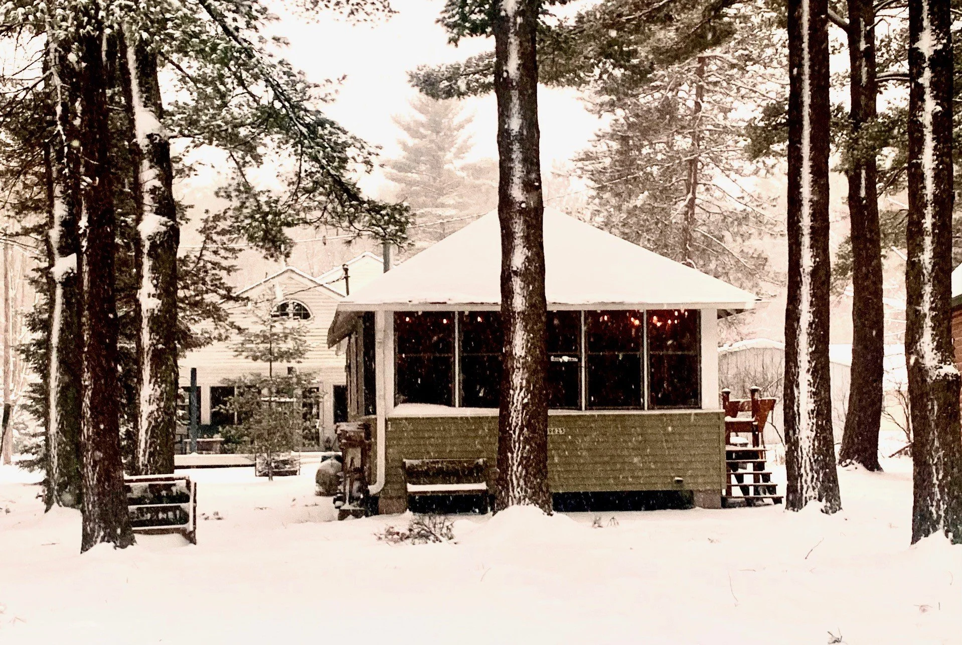 A small house or cabin with a large porch, surrounded by snow-covered trees in a winter landscape.