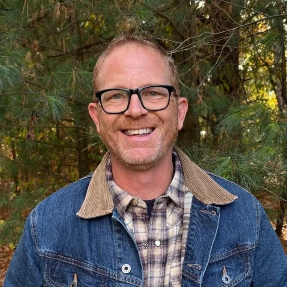 A man with glasses and a beard smiling outdoors in a wooded area, wearing a plaid shirt and denim jacket.