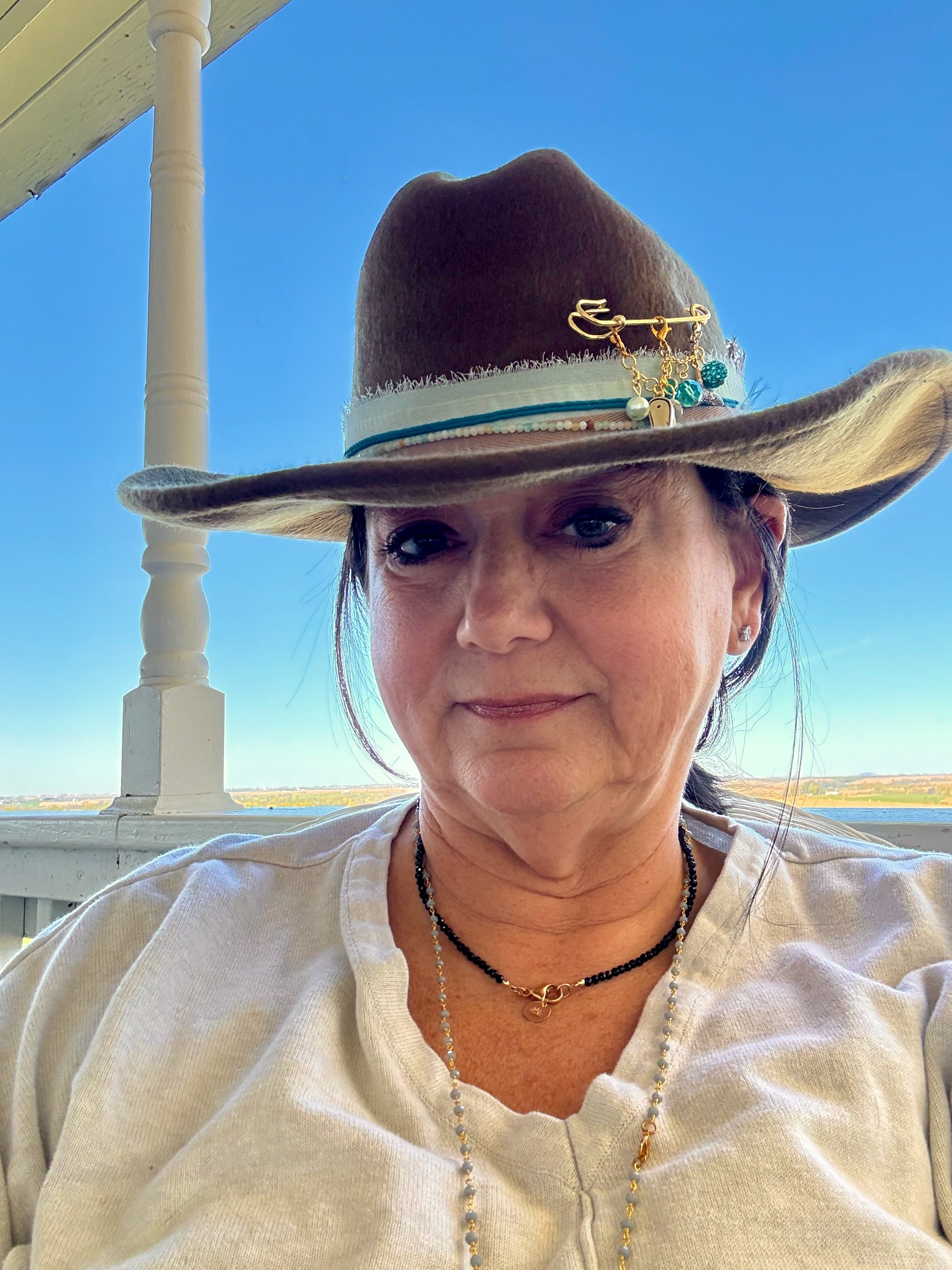A woman wearing a wide-brimmed hat with pins and charms, sitting outdoors on a porch with a clear blue sky and water in the background.