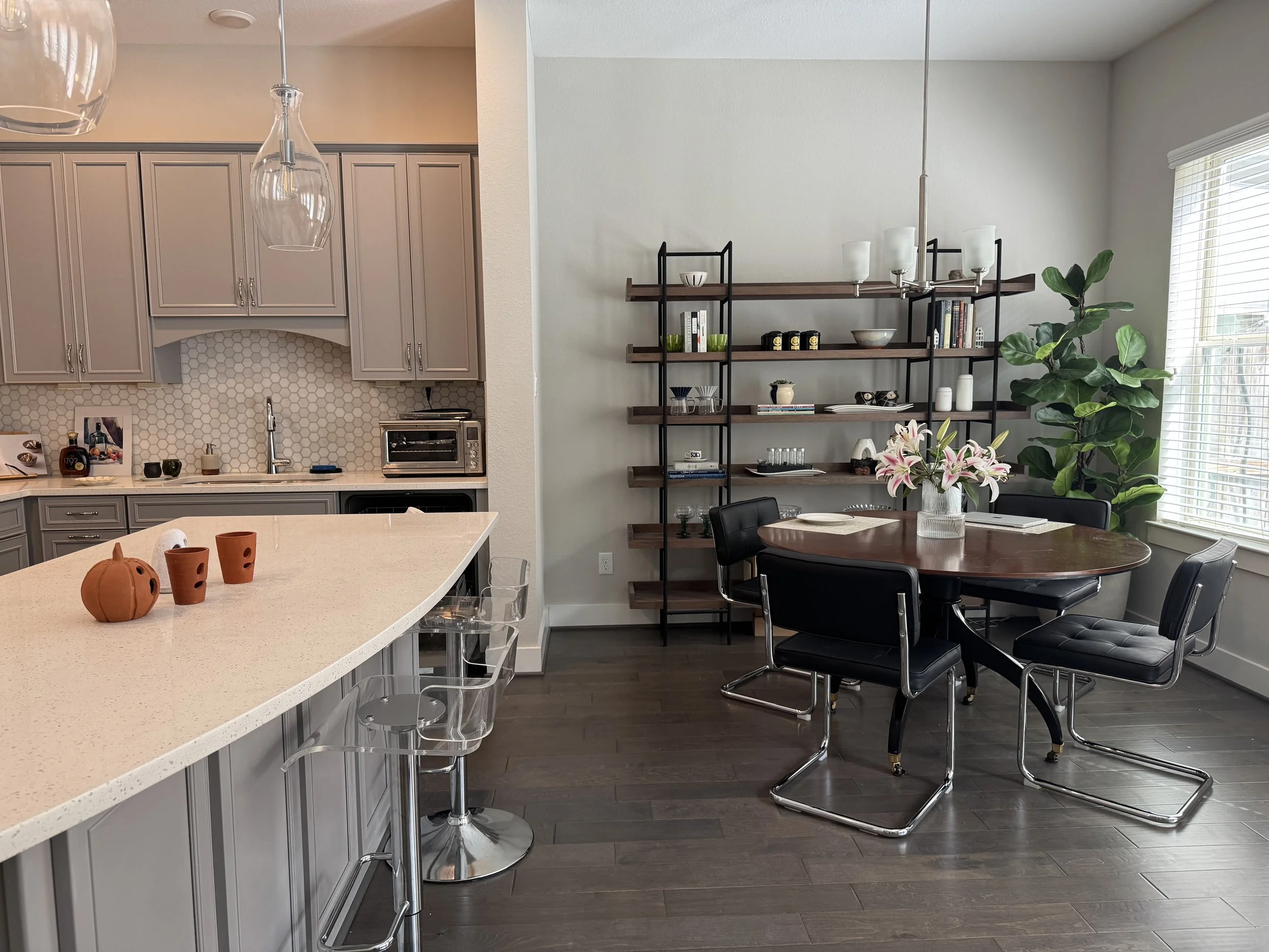 Modern kitchen and dining area with island, gray cabinets, open shelving, black chairs, round wooden table, white flower arrangement, large green plant, window with blinds.