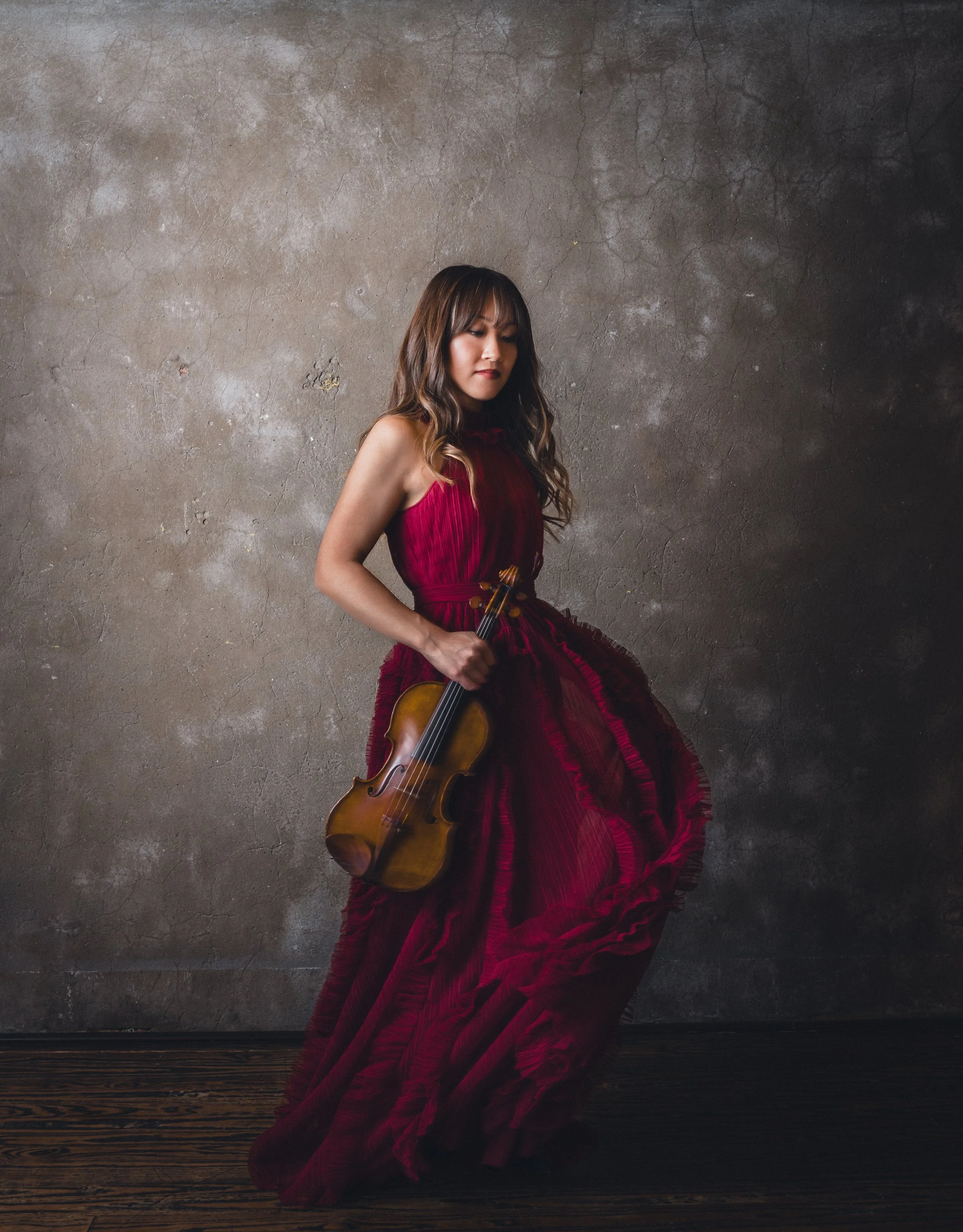 A woman in a flowing red dress holding a violin, standing against a textured concrete wall.