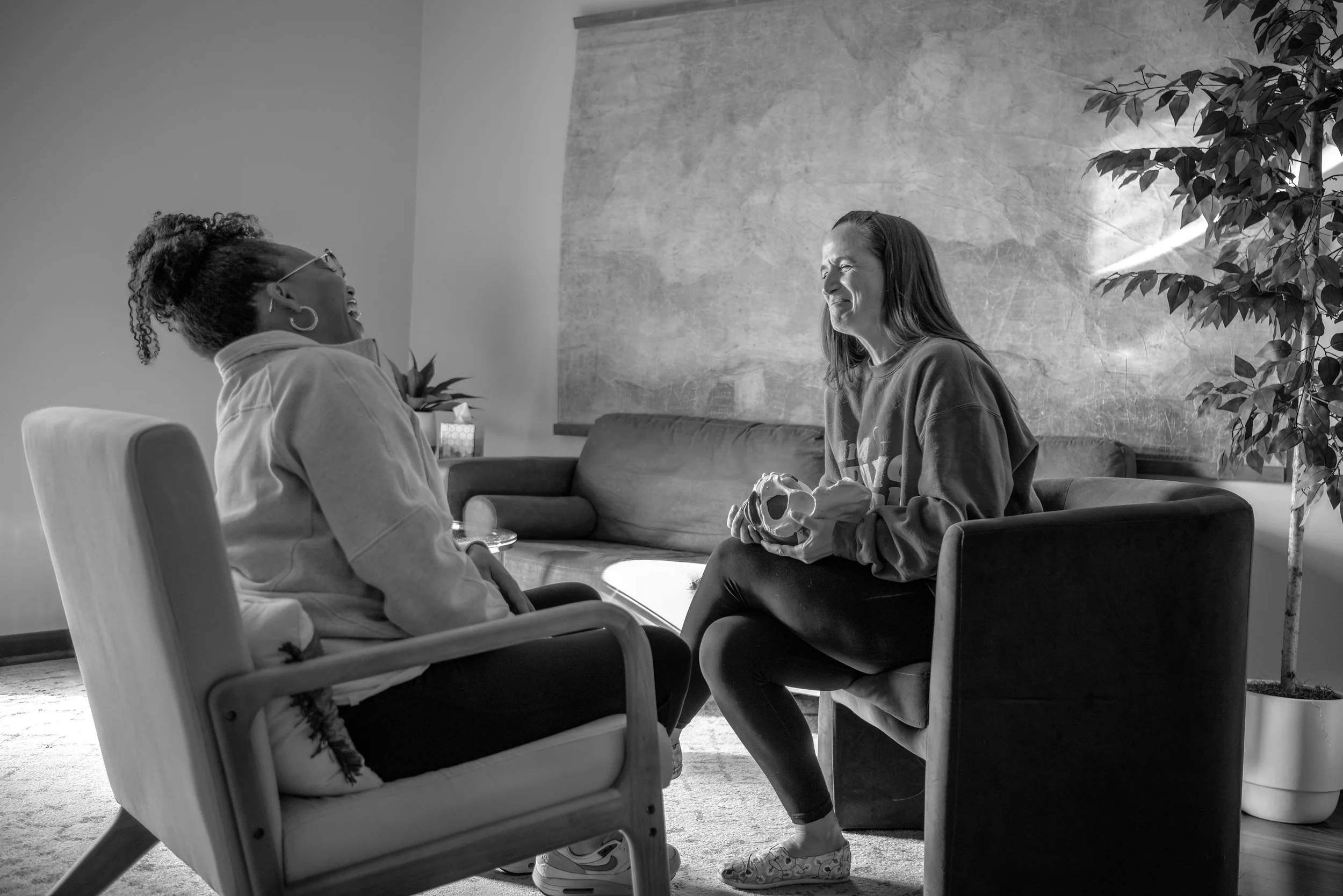Two women sitting and talking in a living room, one holding a skull prop. One woman is wearing glasses and a face mask, the other has long hair and is smiling. There is a potted plant and a large textured wall in the background.