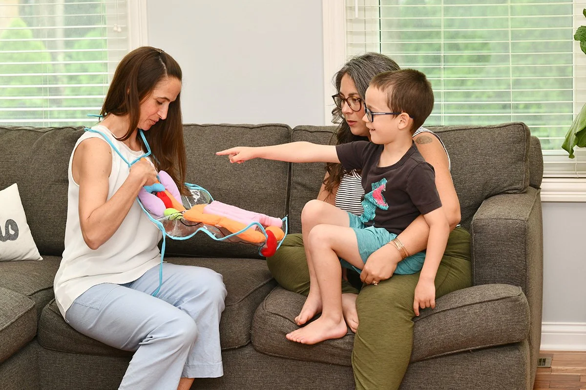 Woman sitting on a sofa holding a colorful sensory toy or plush item while a young boy sitting on a woman's lap points at it, and a woman with gray hair and glasses looks on inside a bright living room with windows.