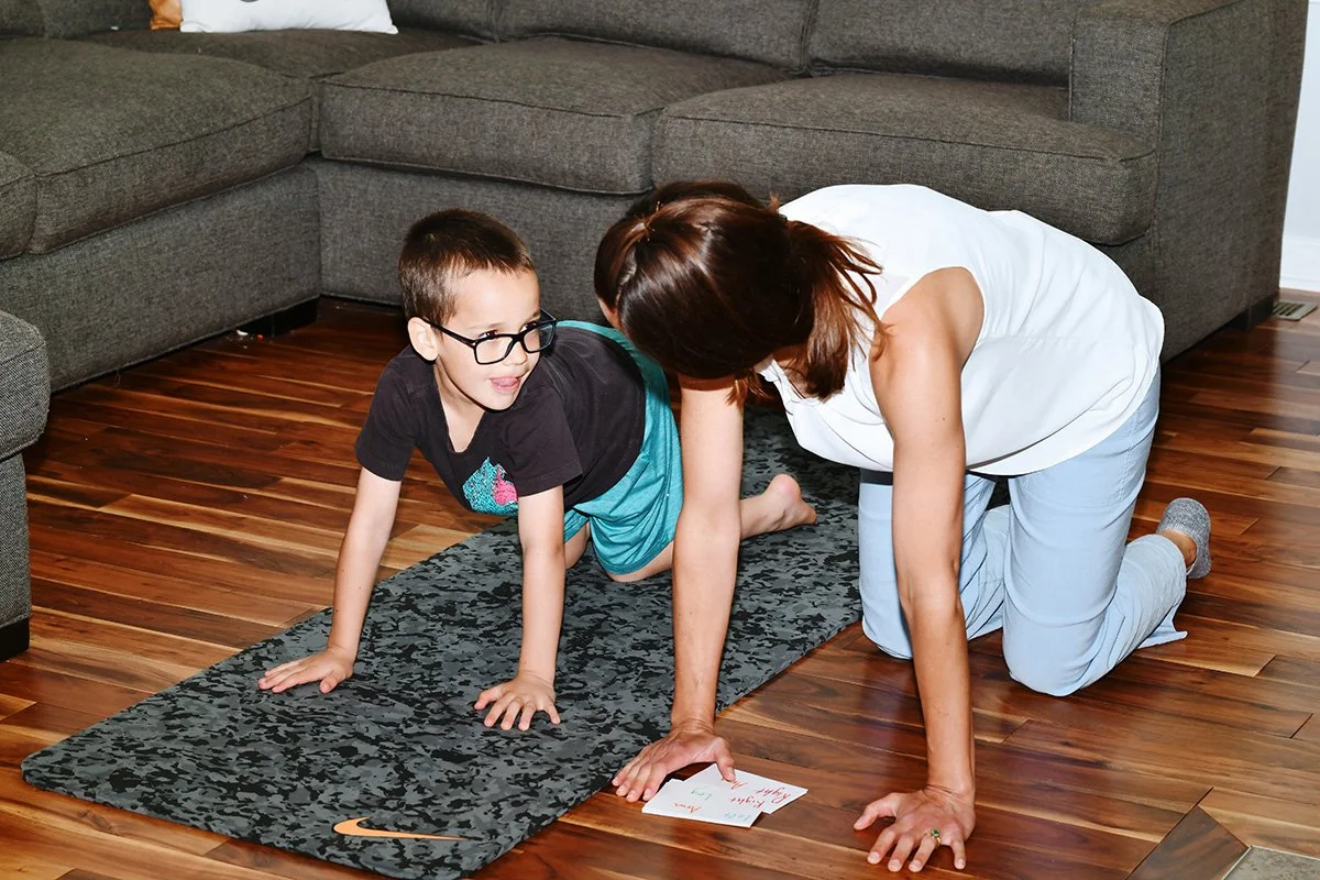 A woman and a young boy doing a yoga pose together on a black patterned yoga mat on a wooden floor in a living room. The woman is on her hands and knees, and the boy is following her lead, looking at her. The woman has brown hair and is wearing a white sleeveless top and light-colored pants. The boy has short brown hair, glasses, a black t-shirt, and blue shorts.