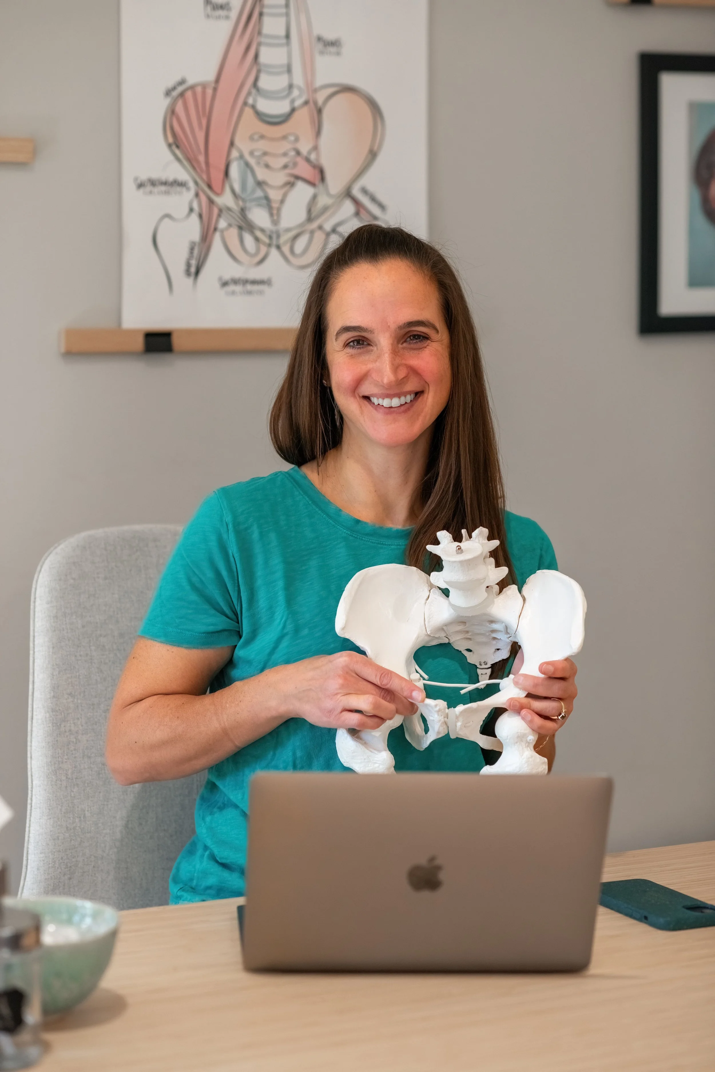 A woman with long brown hair and a turquoise shirt sitting at a desk, holding a pelvis anatomical model, smiling at the camera, with a laptop in front of her and medical diagrams on the wall behind.
