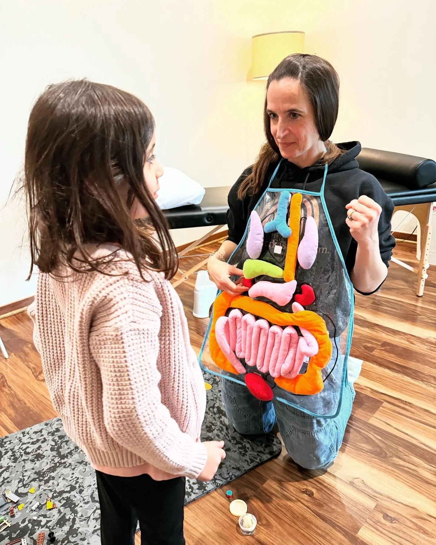 A woman dressed in an apron designed as a colorful human torso with internal organs, kneeling on the floor and talking to a young girl in a pink sweater, inside a room with wooden floors and a massage table.