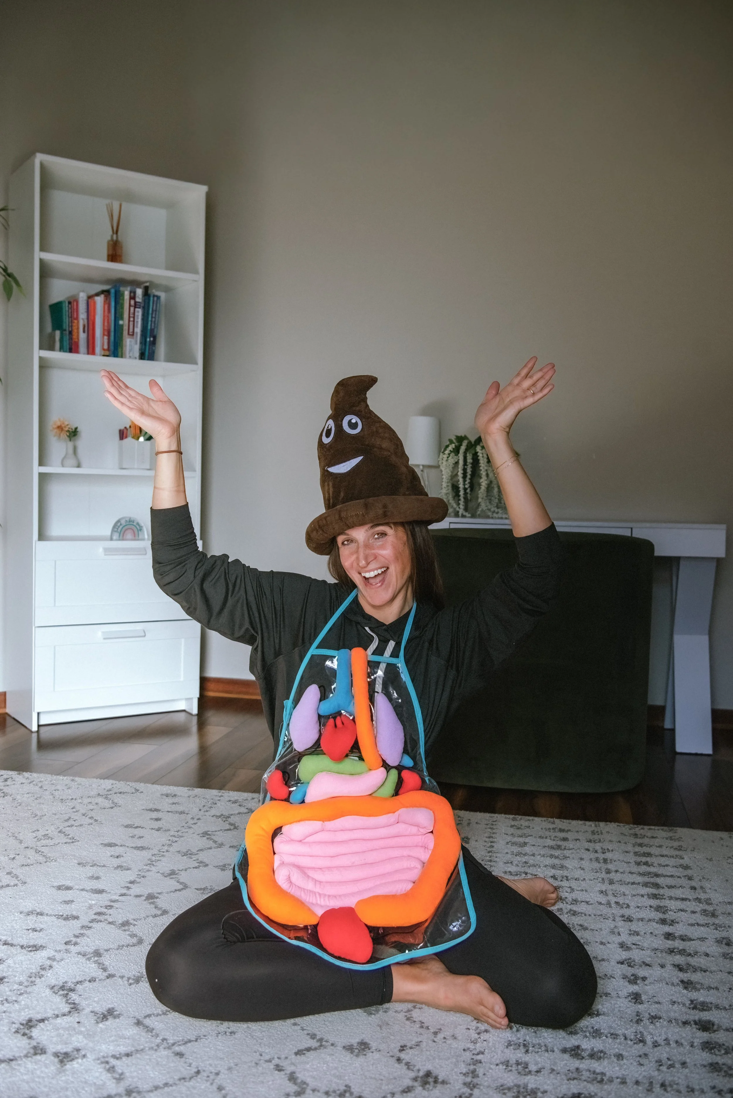 A woman sitting cross-legged on a carpeted floor, smiling and raising her arms, wearing a plush poop emoji hat with a smiling face and a colorful plush intestines apron.