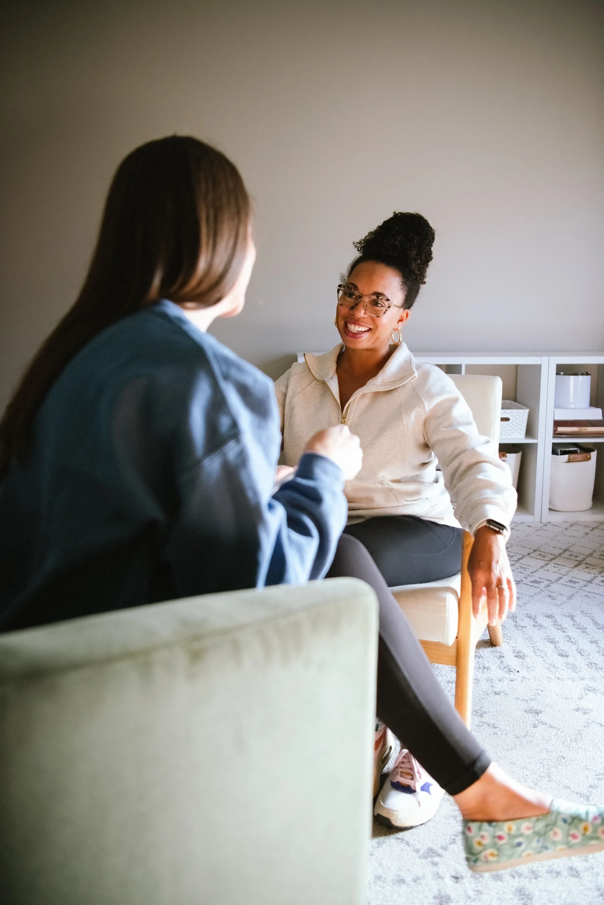 Two women having a conversation in a cozy indoor setting, one with curly hair and glasses smiling, the other woman with straight hair whose back is facing the camera.