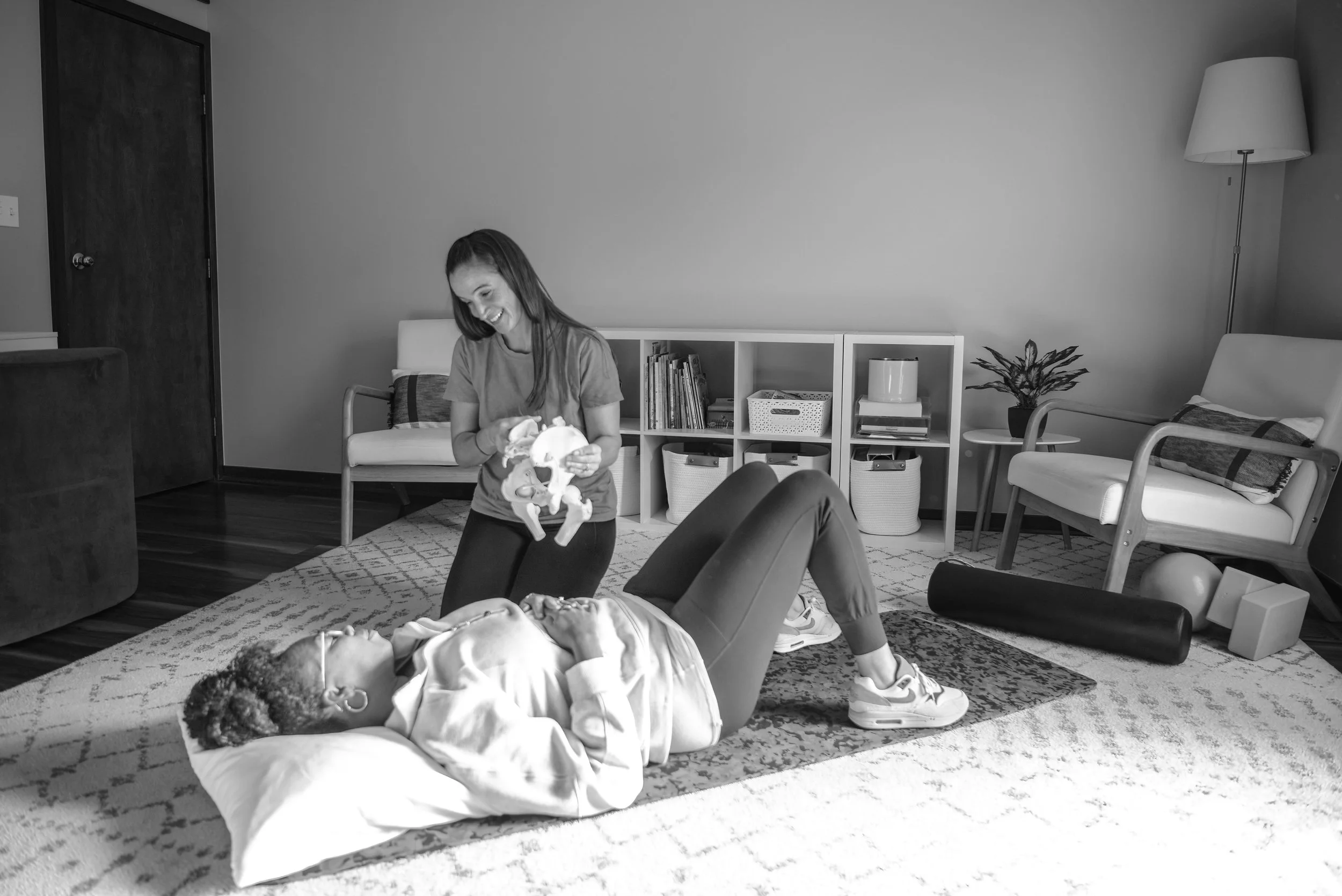A woman lying on a bed doing pelvic exercises with a weighted ball held between her legs, while another woman is standing nearby holding a stuffed animal and smiling, in a living room.