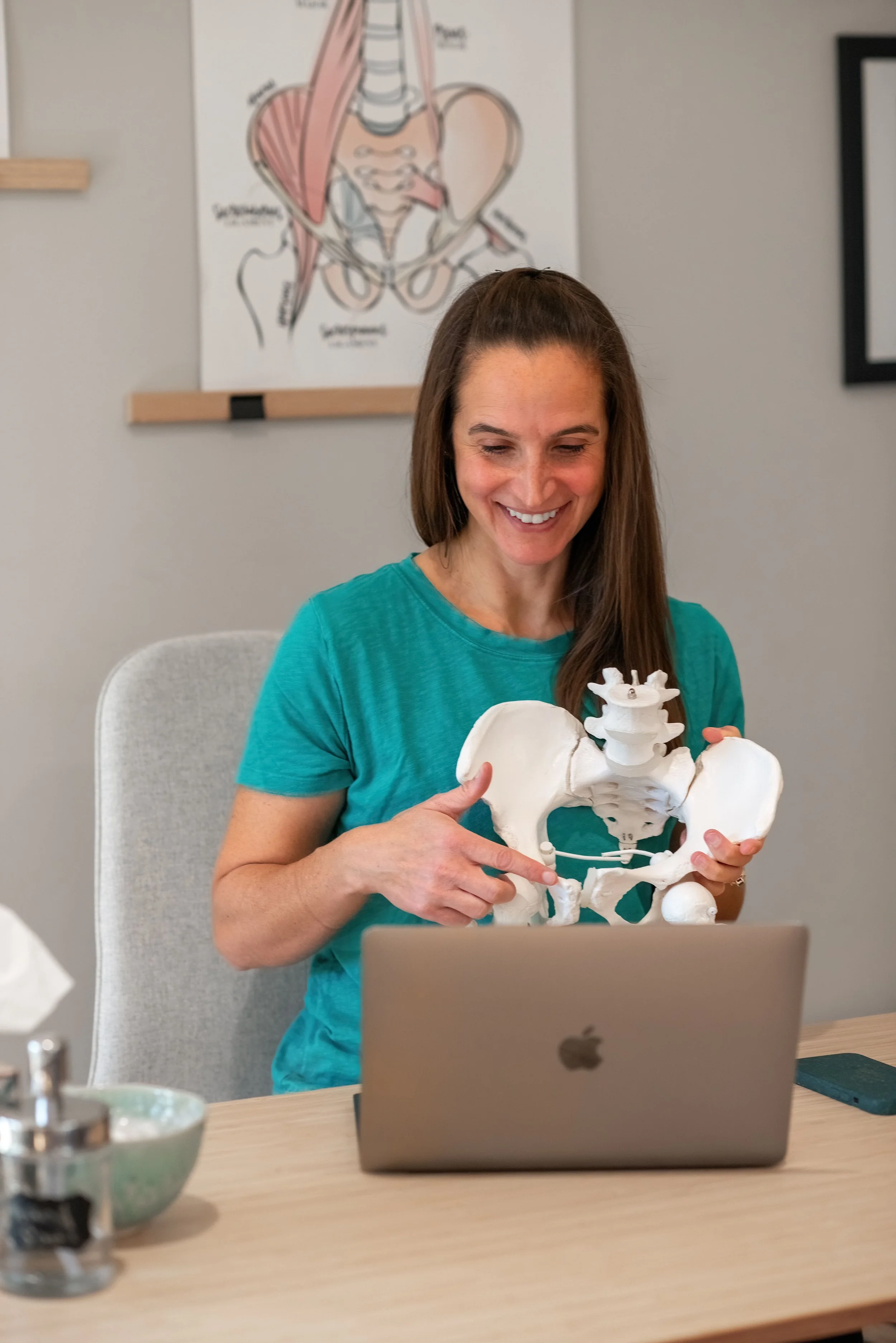 A woman with long brown hair smiling and pointing at a white pelvic bone model she is holding, sitting at a desk with a laptop in front of her.