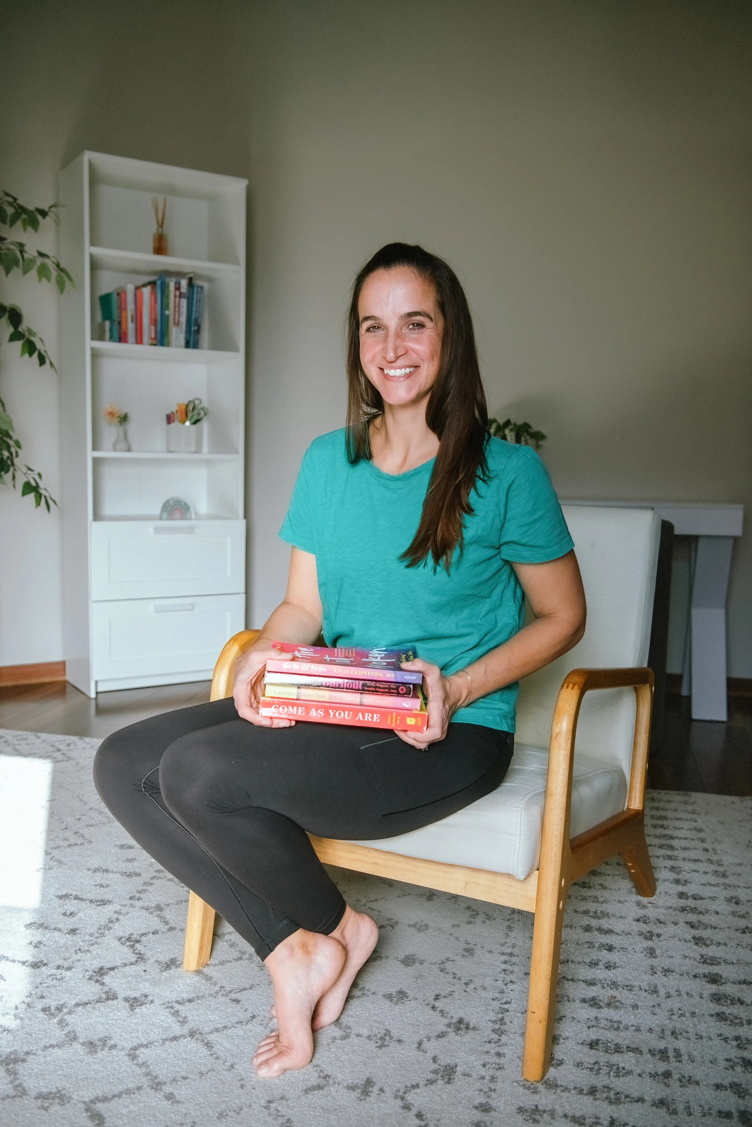 A woman with long dark hair, wearing a teal T-shirt and black pants, sits on a wooden chair without shoes, holding a stack of colorful books and smiling at the camera in a living room.