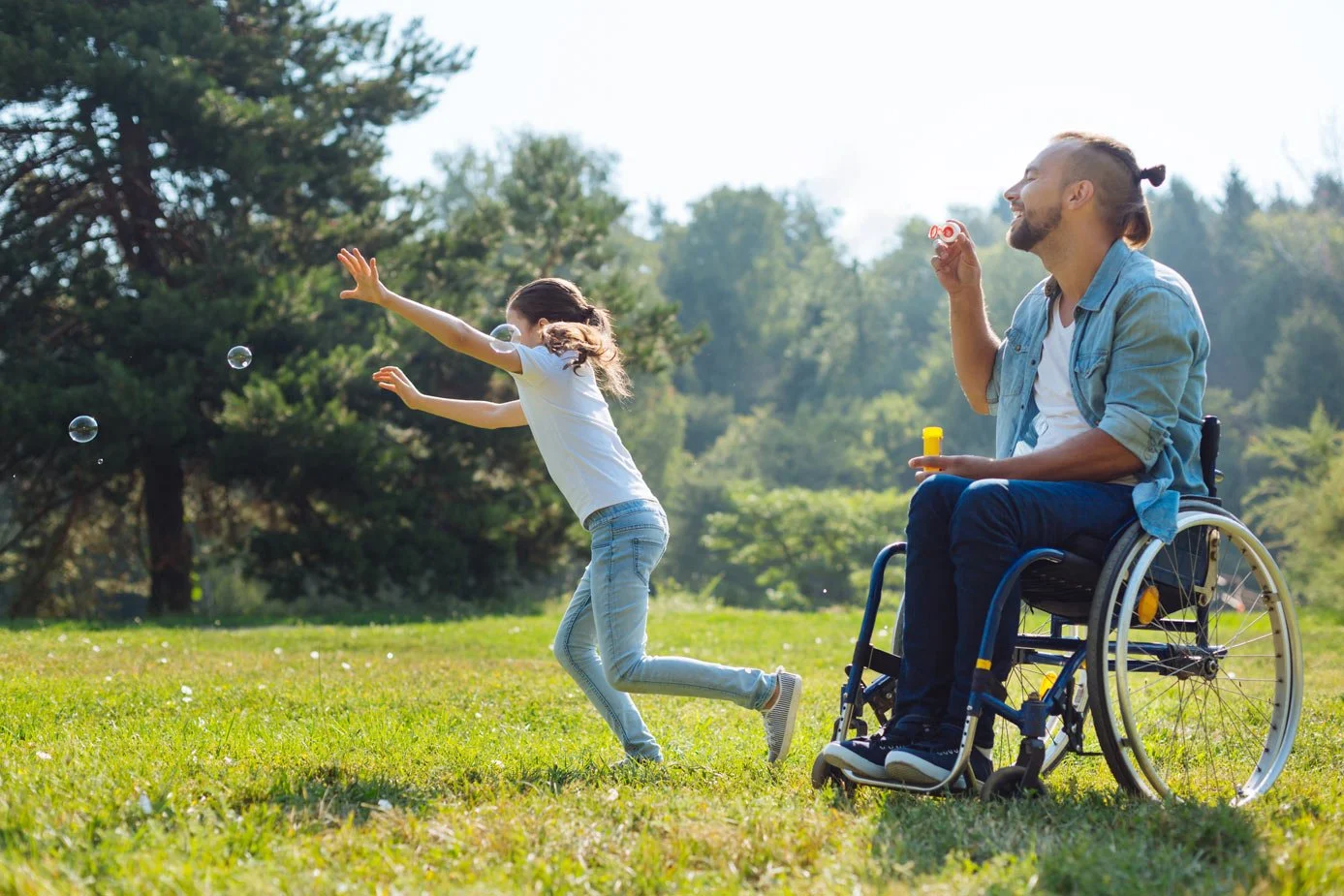 A young girl and a man in a wheelchair are playing with soap bubbles outdoors on a sunny day in a park with green grass and trees in the background.