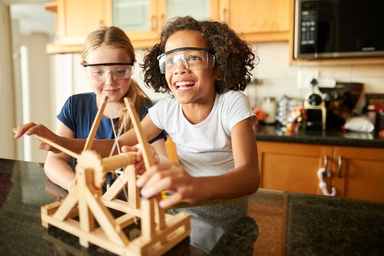 Two young girls, wearing safety goggles, are smiling and playing with a wooden catapult model on a kitchen counter.