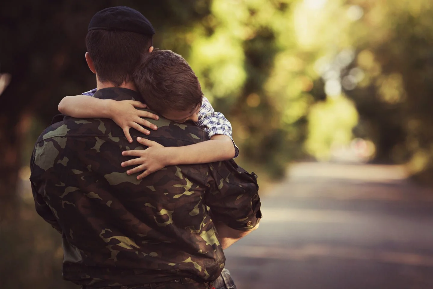 A soldier in camouflage uniform holding a boy on his shoulder, hugging him tightly outdoors on a sunny day.