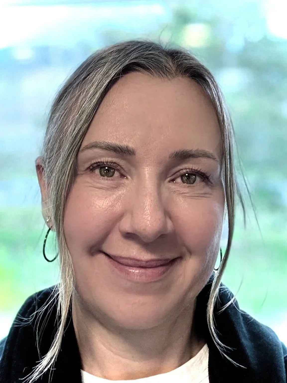 A woman with blonde hair, wearing hoop earrings and a black top, smiling in front of a window with a blurred outdoor background.