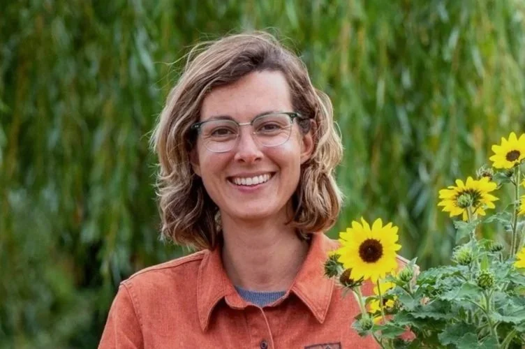 A woman with curly, shoulder-length brown hair, wearing glasses and an orange shirt, smiling outdoors in a sunflower field.