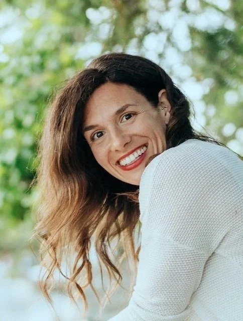 A woman smiling outdoors with trees in the background.