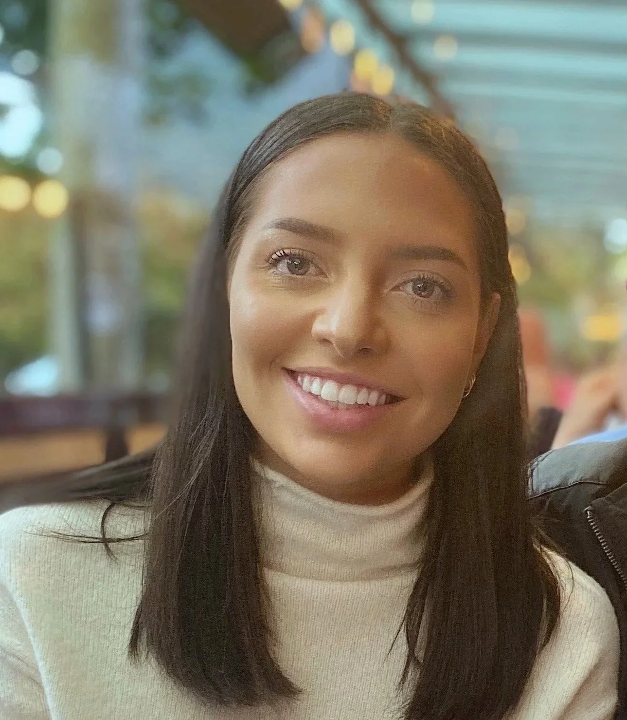 A young woman with long dark hair smiling at the camera, sitting indoors with a blurred background of colorful lights and windows.