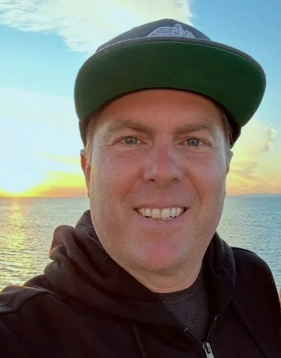A man smiling outdoors at sunset near a body of water, wearing a black hoodie and a baseball cap.