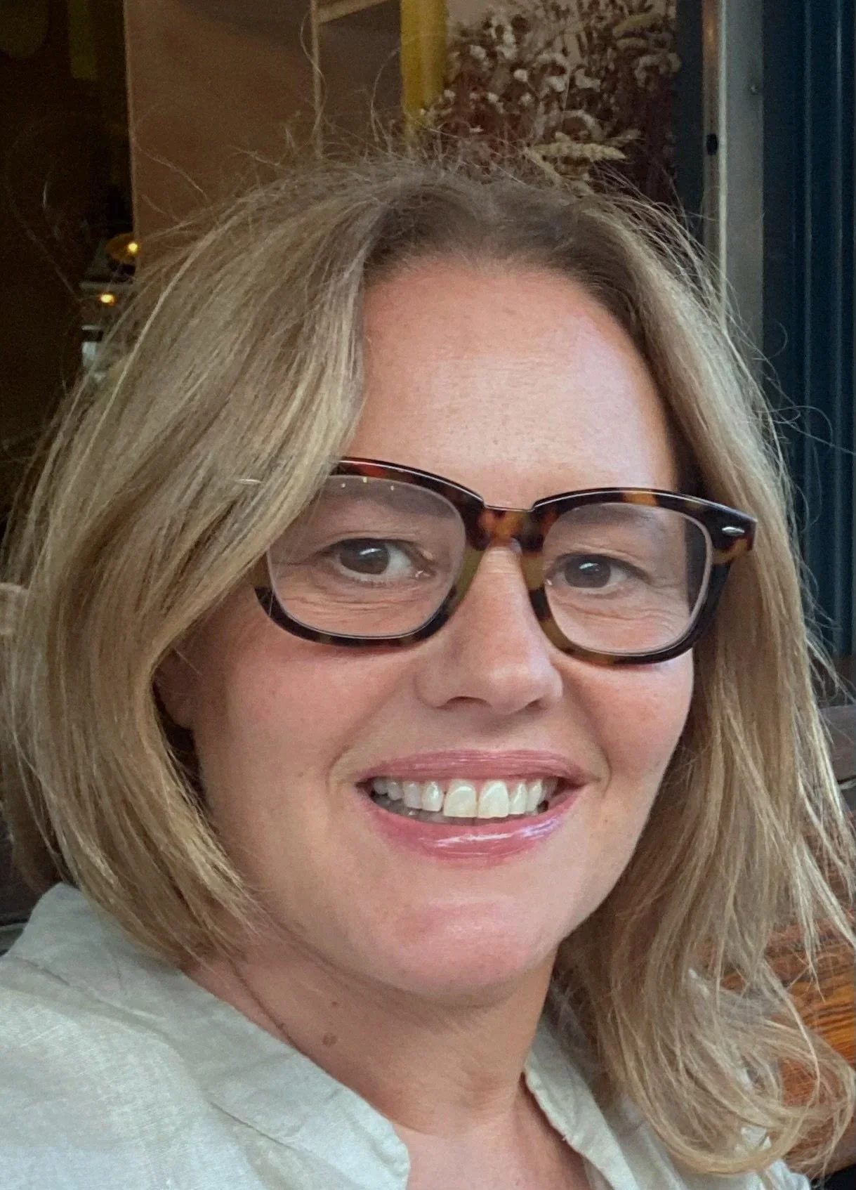 A woman with blonde hair wearing tortoiseshell glasses, smiling, in a cozy indoor setting.