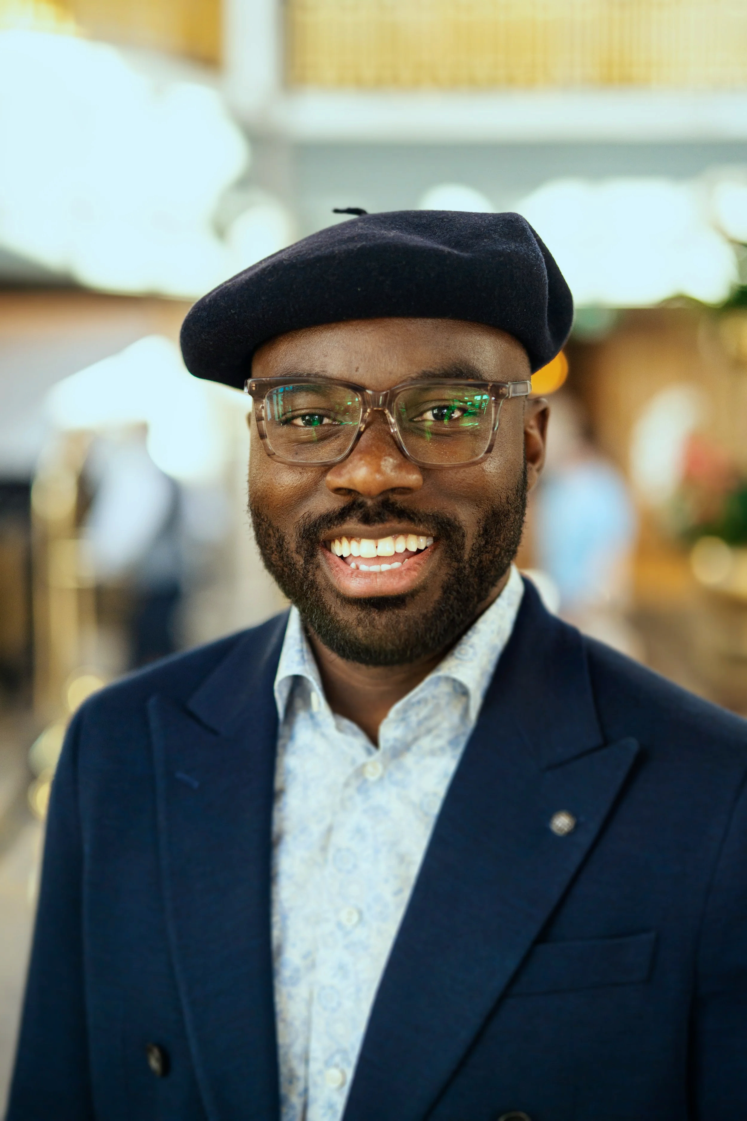A smiling man wearing glasses, a black beret, a white patterned shirt, and a navy blazer in an indoor setting.