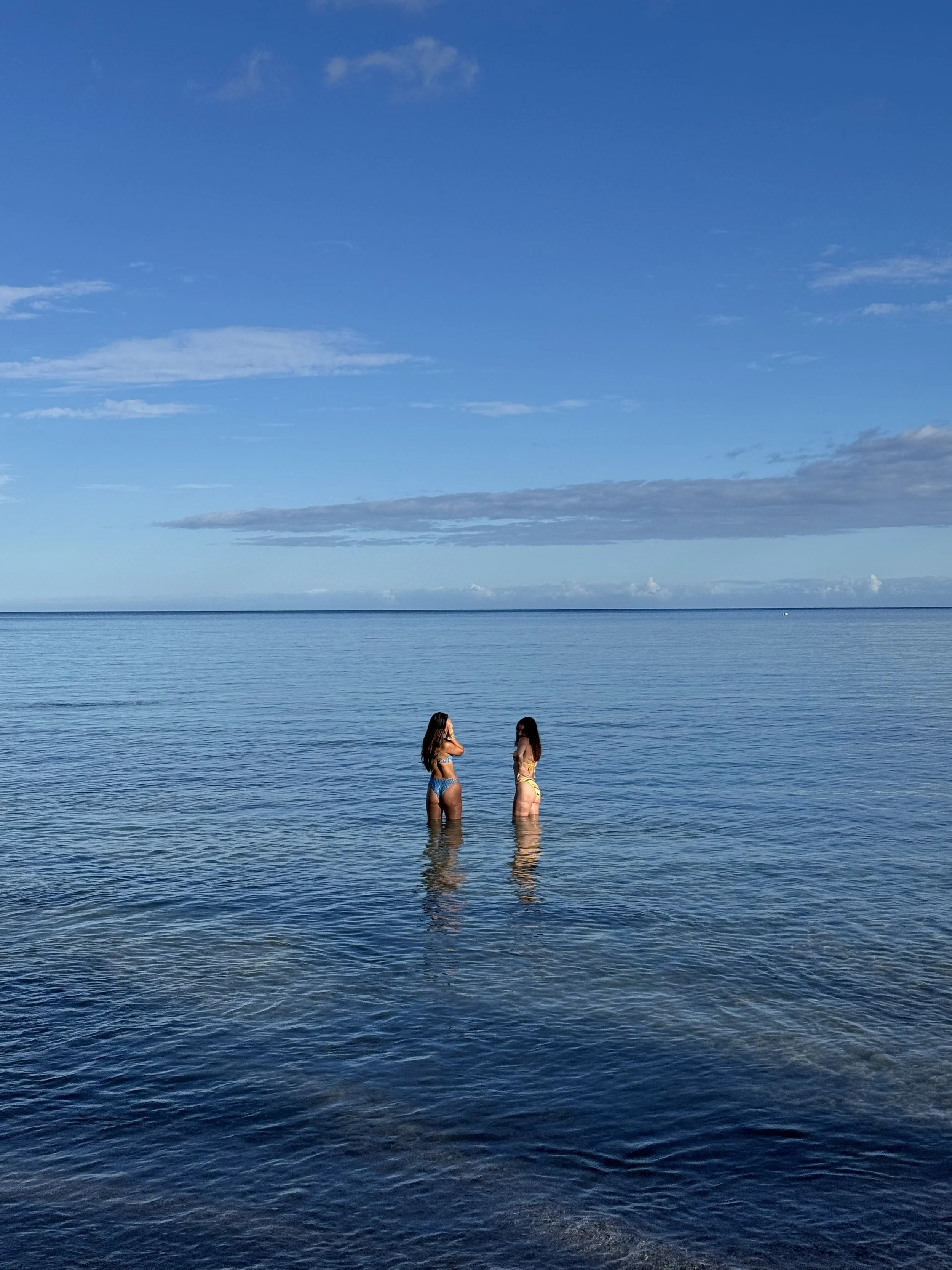 Two women in swimsuits standing waist-deep in calm ocean water under a blue sky with scattered clouds.