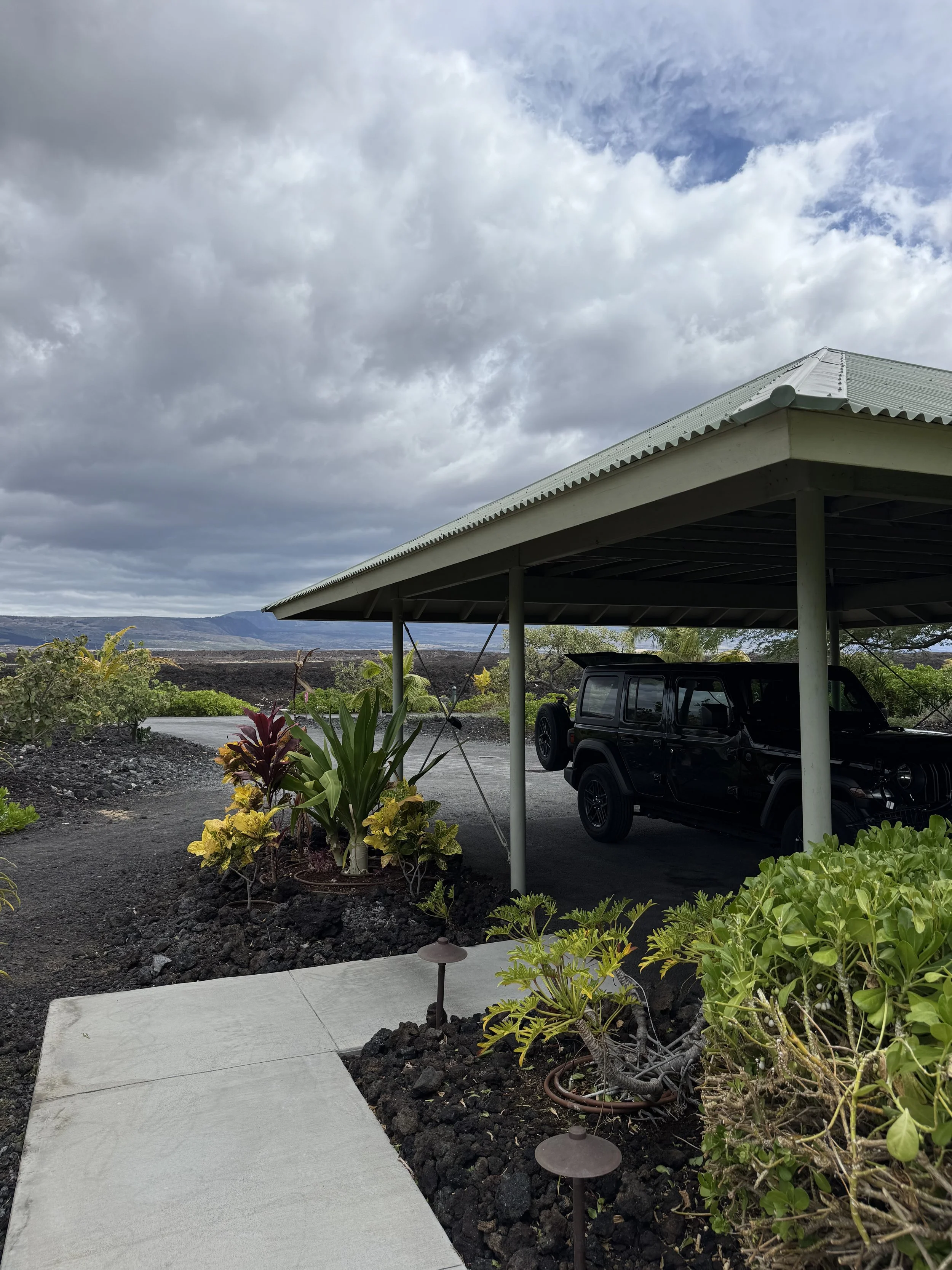 A black SUV parked under a metal-roofed carport with a garden and dirt road nearby, under a cloudy sky.