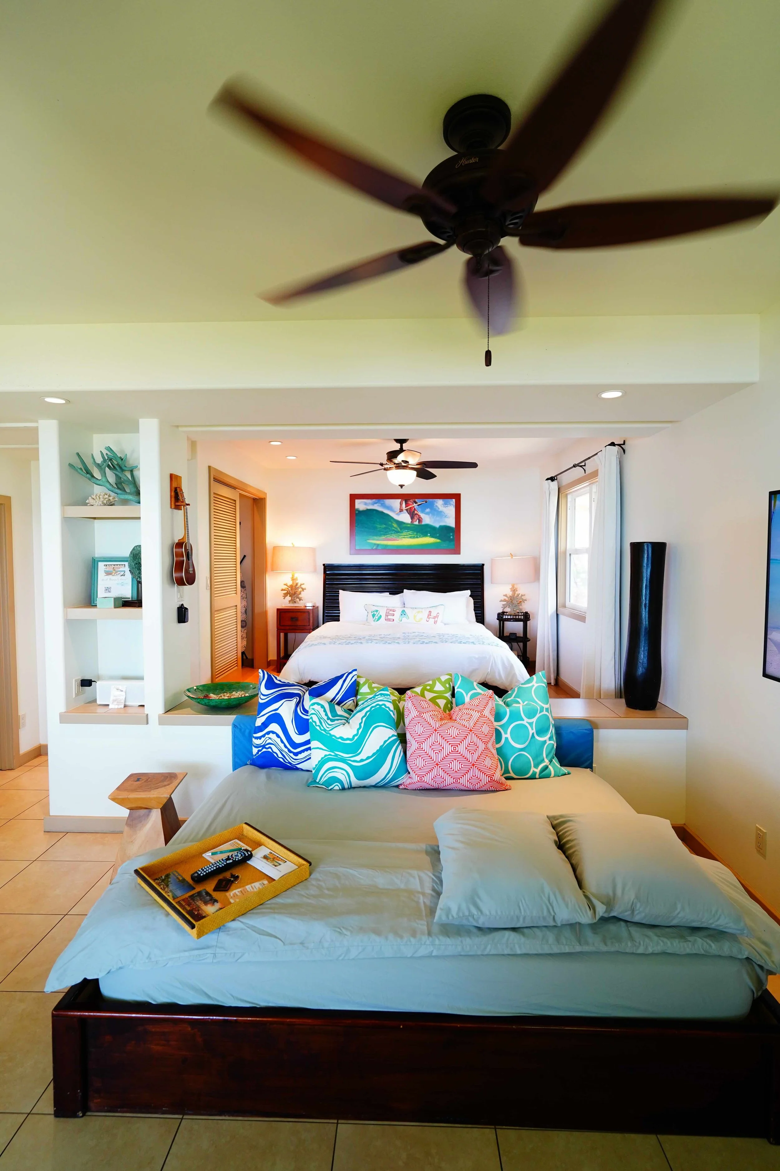 Hotel room with a bed, colorful pillows, and a view of the bedroom and living area, featuring ceiling fans, a dresser, and decor with beach-themed accents.