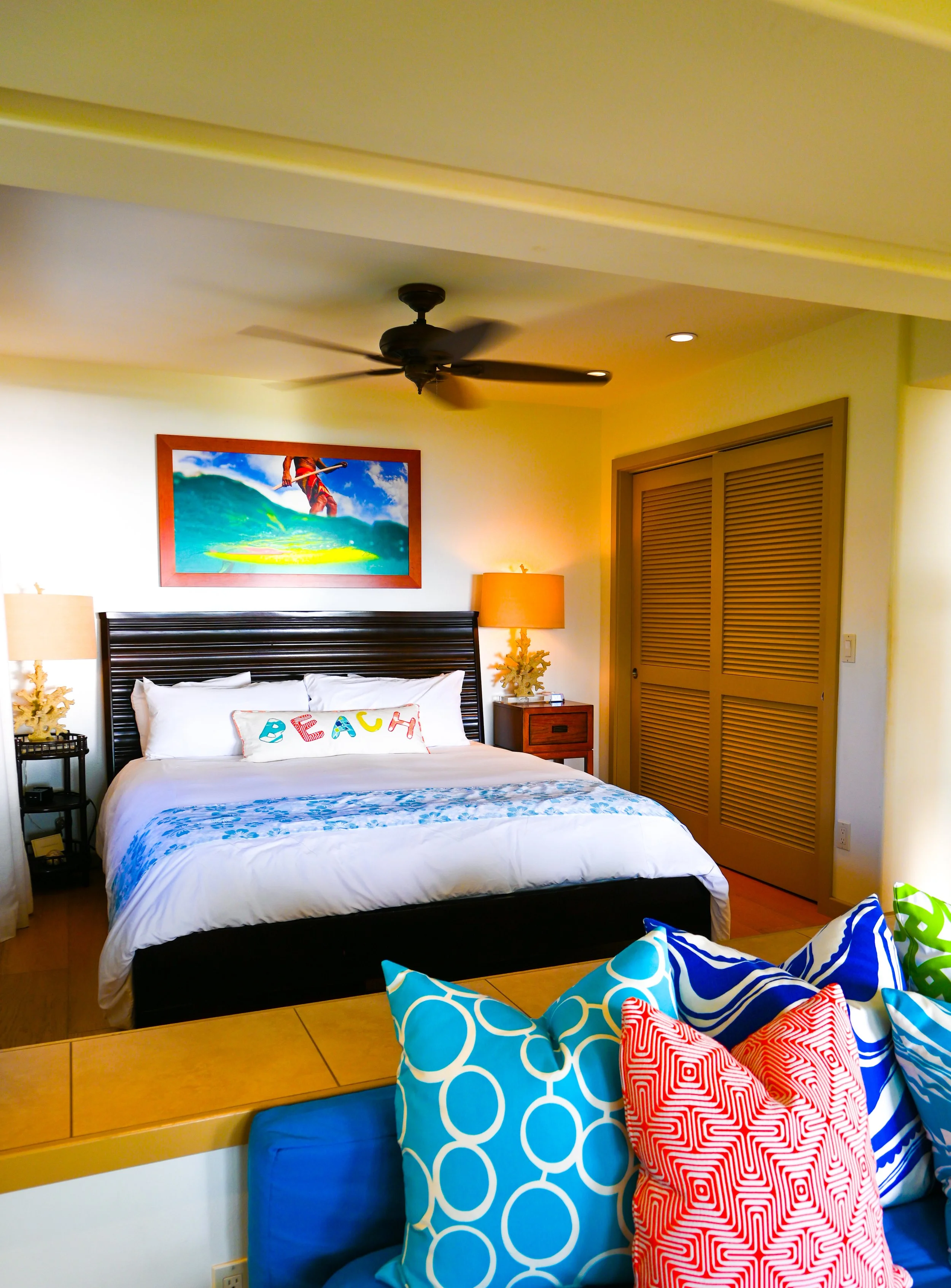A bedroom with a bed that has white bedding and a pillow spelling out 'PEACE'. There are two nightstands with lamps, a colorful framed artwork of a surfer riding a wave above the bed, and a ceiling fan. Colorful pillows are in the foreground.