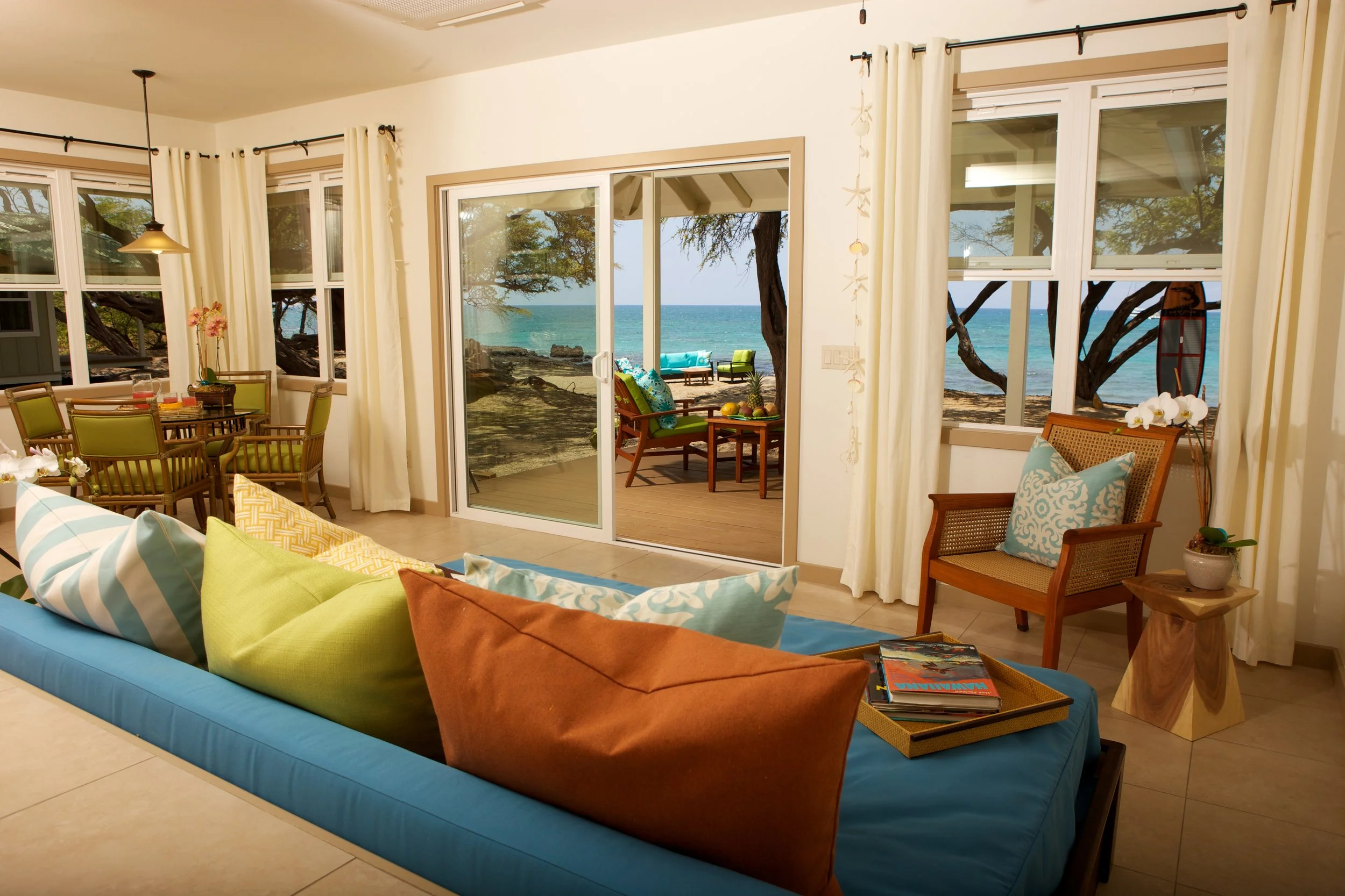 Living room with sliding glass doors leading to outdoor patio overlooking the ocean. Inside, there are colorful cushions on a blue sofa, a wooden armchair with a blue pillow, a small side table with a potted orchid, and a dining area with a large table and green chairs.