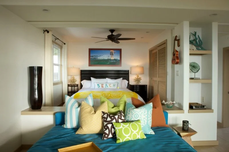 Beach-themed bedroom with a bed, multiple colorful pillows, artwork of a beach scene, and coastal decor.