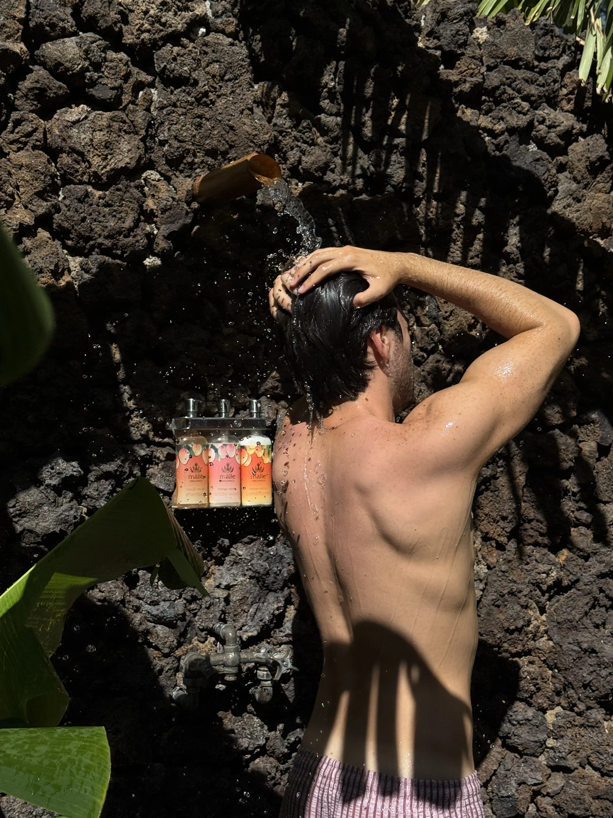 A man with dark hair is taking a shower outdoors against a dark, rocky wall. He is standing with his back to the camera, using one hand to wash his hair. There are green plants nearby, and three bottles of shampoo or body wash are mounted on the wall