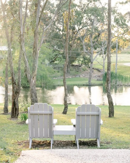 Two white Adirondack chairs facing a lake surrounded by trees.