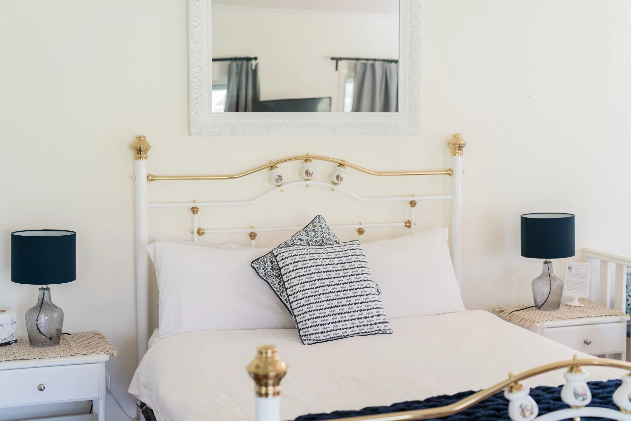 A neatly made bed with white bedding, two pillows with decorative cases, and a vintage-style metal headboard with gold accents. There are navy blue lamps on each side on white nightstands with beige woven covers, and a mirror and window with curtains in the background.