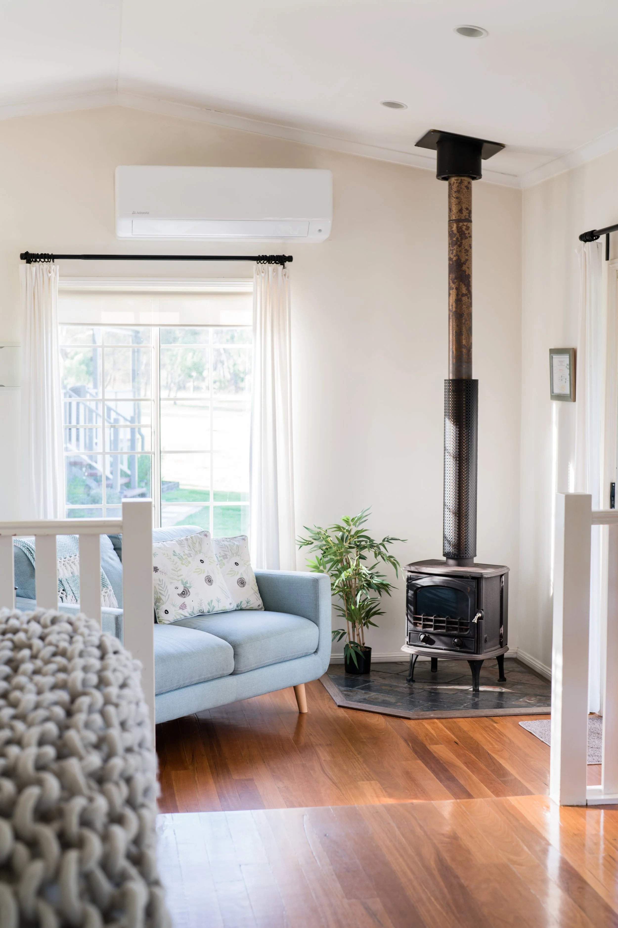 Living room with a light blue sofa, decorative pillows, a wood stove with a rusty flue pipe, a plant, white curtains, a window, and hardwood floors.