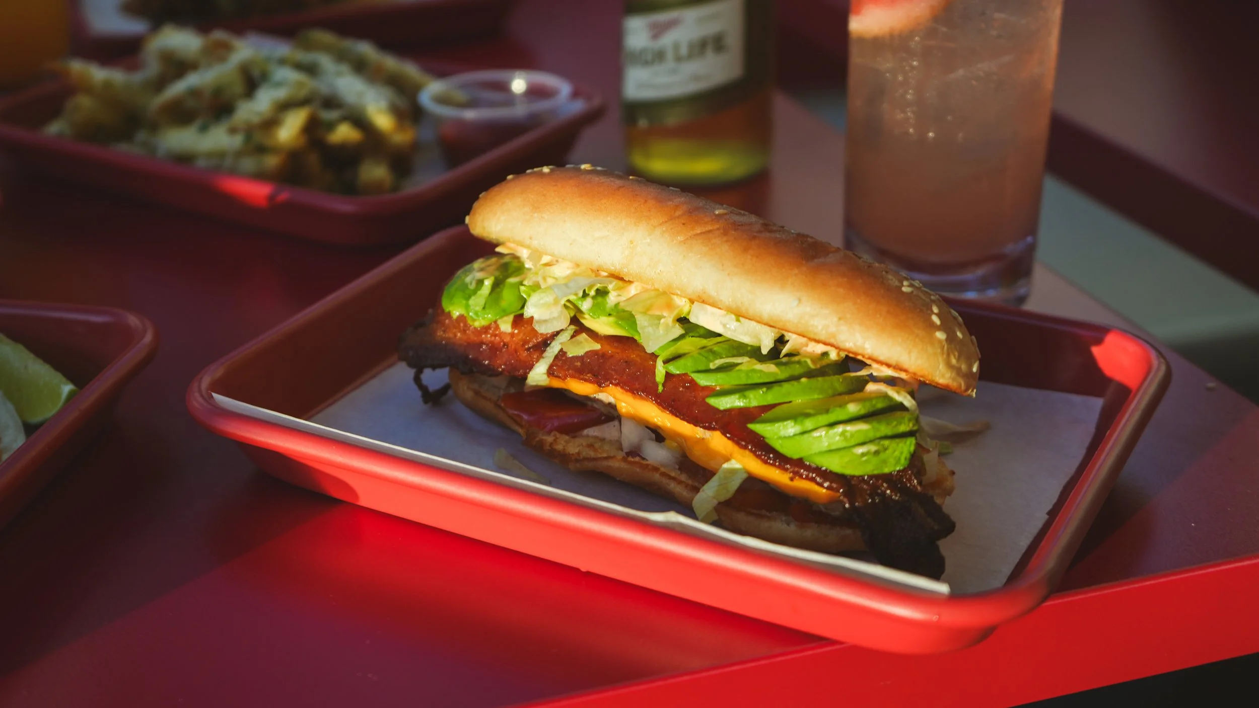 A sandwich with fried chicken, lettuce, avocado, and cheese on a sesame seed bun, served on a red tray at quik dog.
