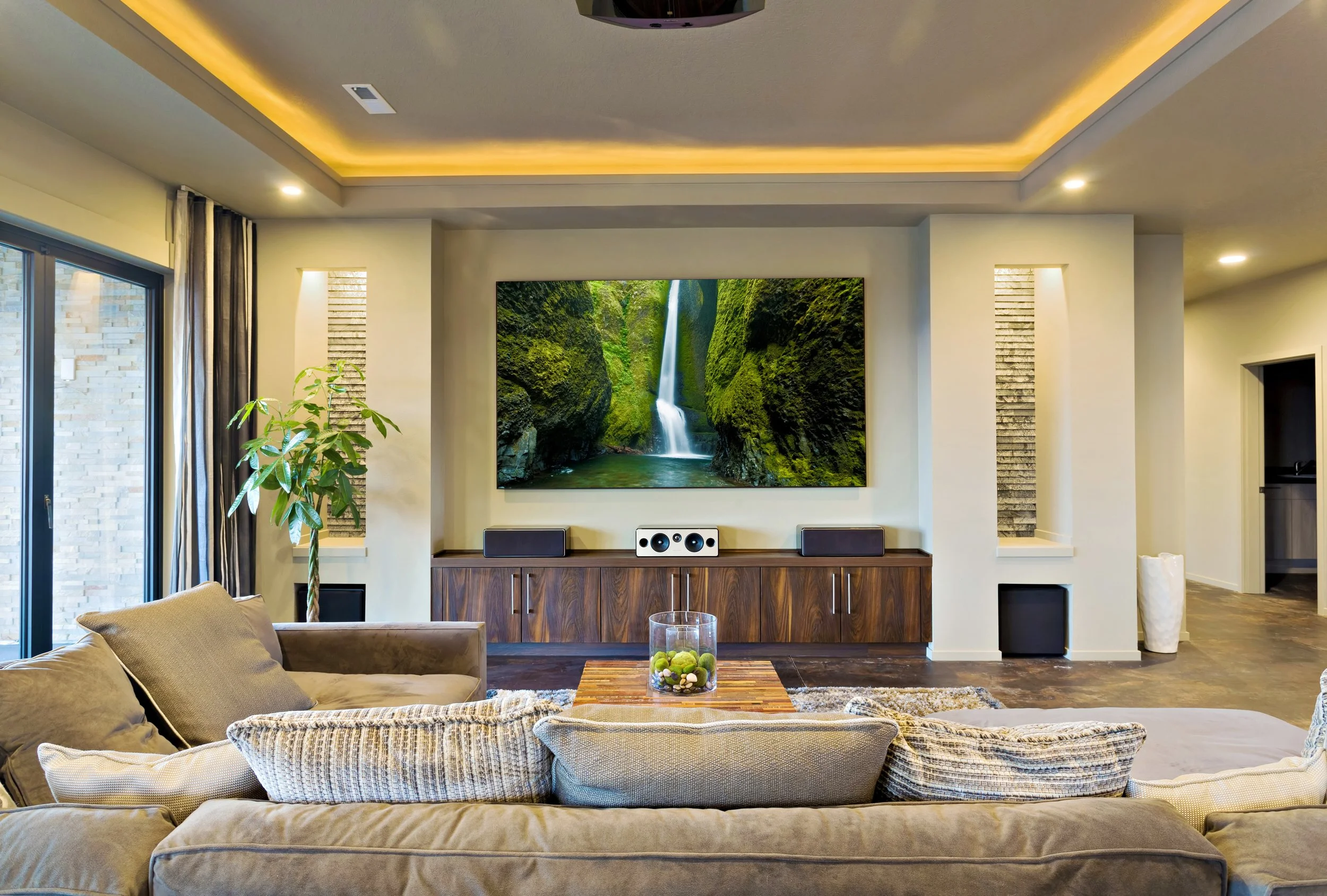Living room with beige couches, a glass-topped coffee table, large window with curtains, a wooden media console with a flat-screen TV displaying a waterfall scene, and built-in wall lighting.
