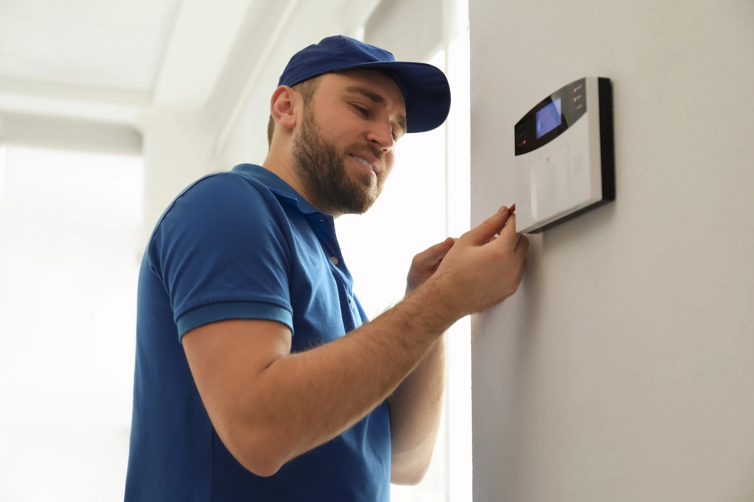A delivery person in a blue uniform and cap adjusting a security system panel on a white wall.